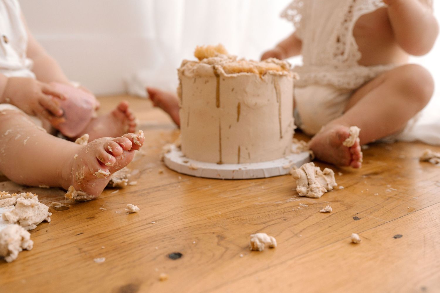Two babies sit on a wooden floor around a smashed first birthday cake during a cake smash photo session.