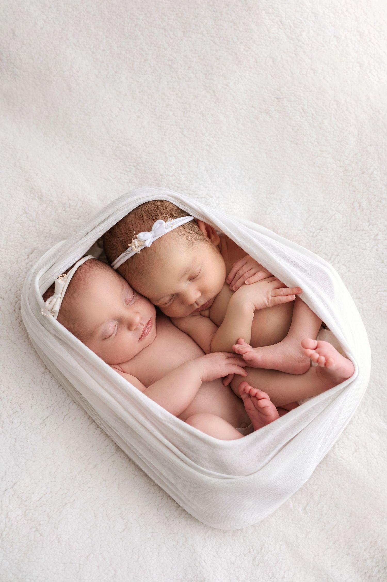 Two sleeping newborn babies nestled together in a white cushioned basket wearing delicate headbands.