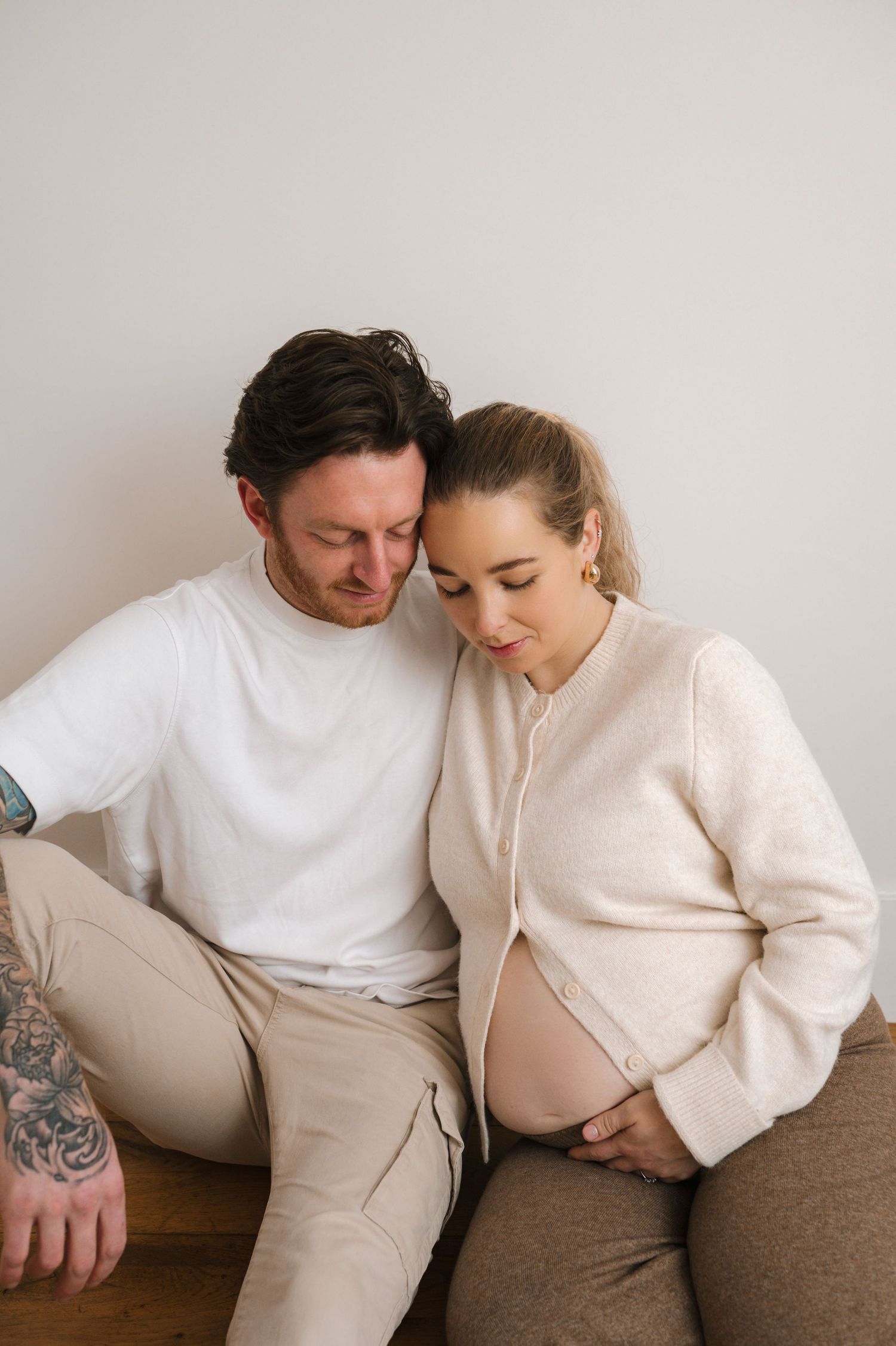 A couple sits together, the man leaning close to his pregnant partner as she cradles her baby bump tenderly.