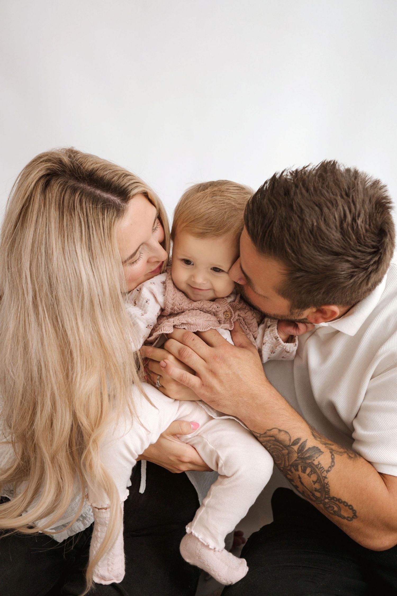 Loving parents share tender moment with their baby in white outfit against neutral background.