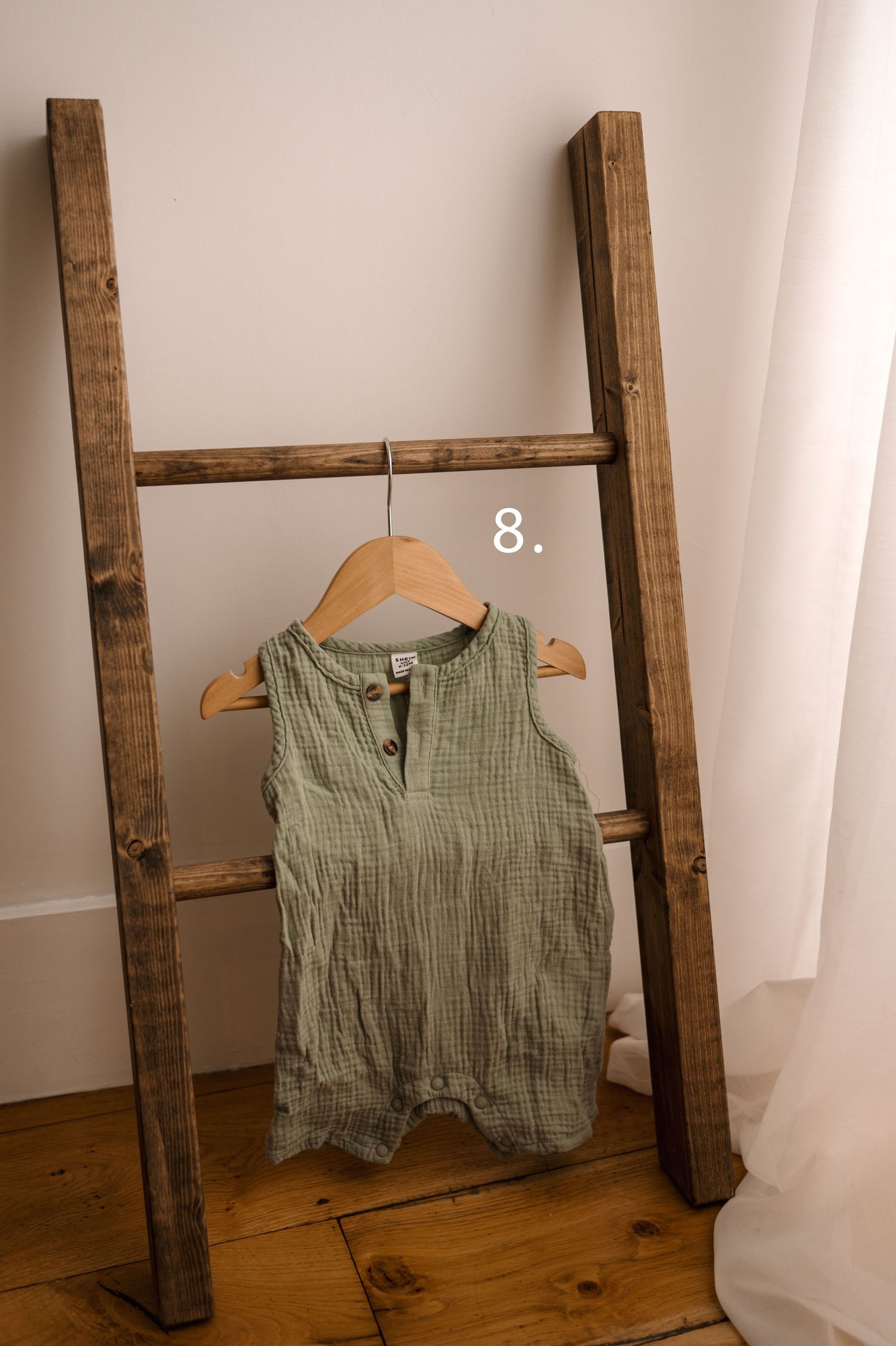 Sage green textured baby romper displayed on wooden hanger against rustic ladder and white fabric background.