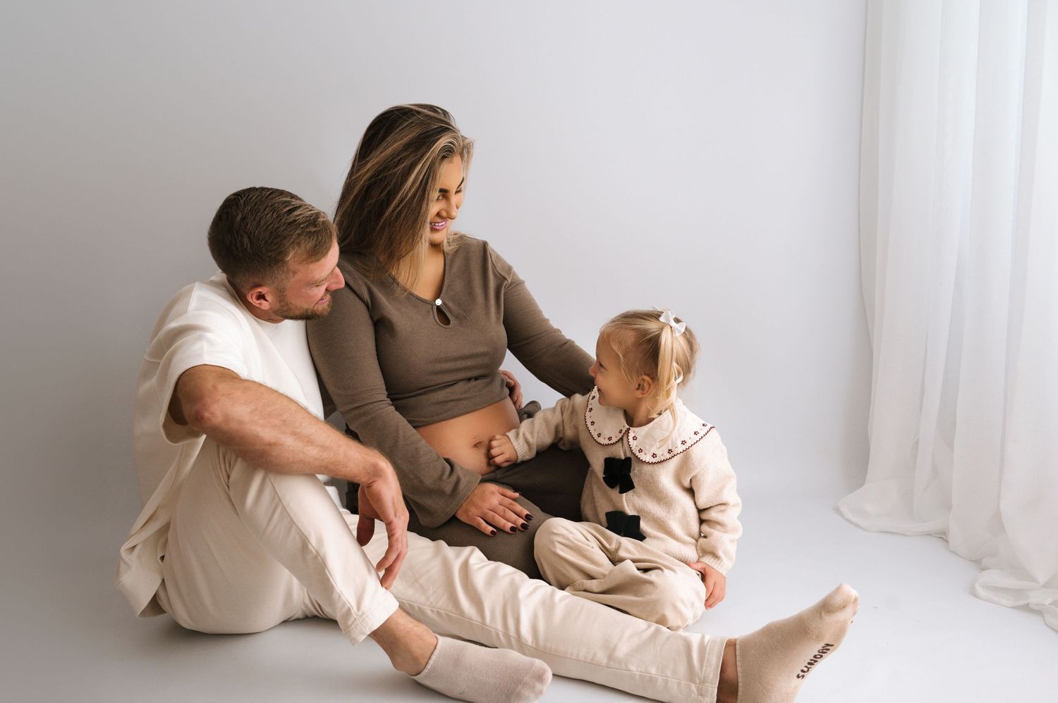 Family sitting together on floor in white room wearing neutral colors with small dog.