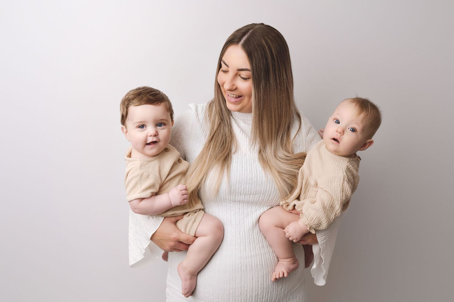 Mother in white dress holds two babies in matching cream outfits against neutral backdrop for family portrait.
