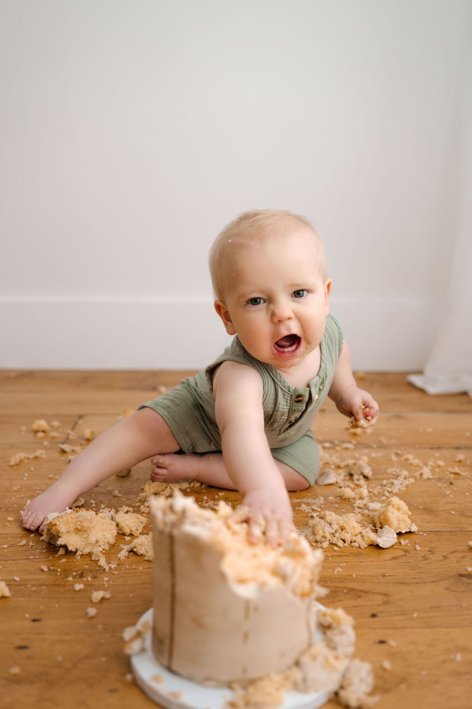 Baby in green romper smashing first birthday cake on wooden floor, covered in crumbs, mouth open with joy.
