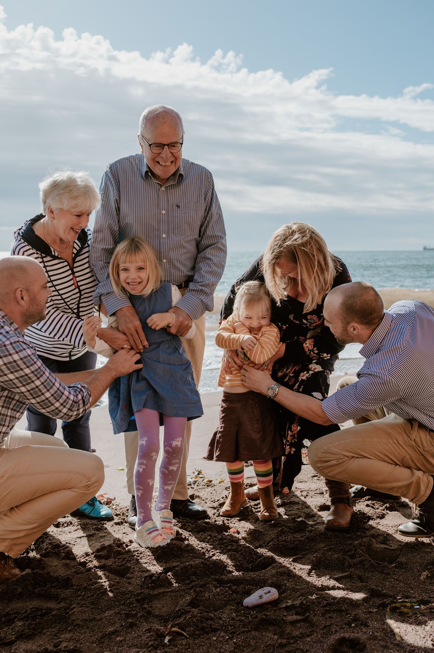 Multi-generational family gathering on a beach, grandparents and parents smiling at two young girls playing in the sand.