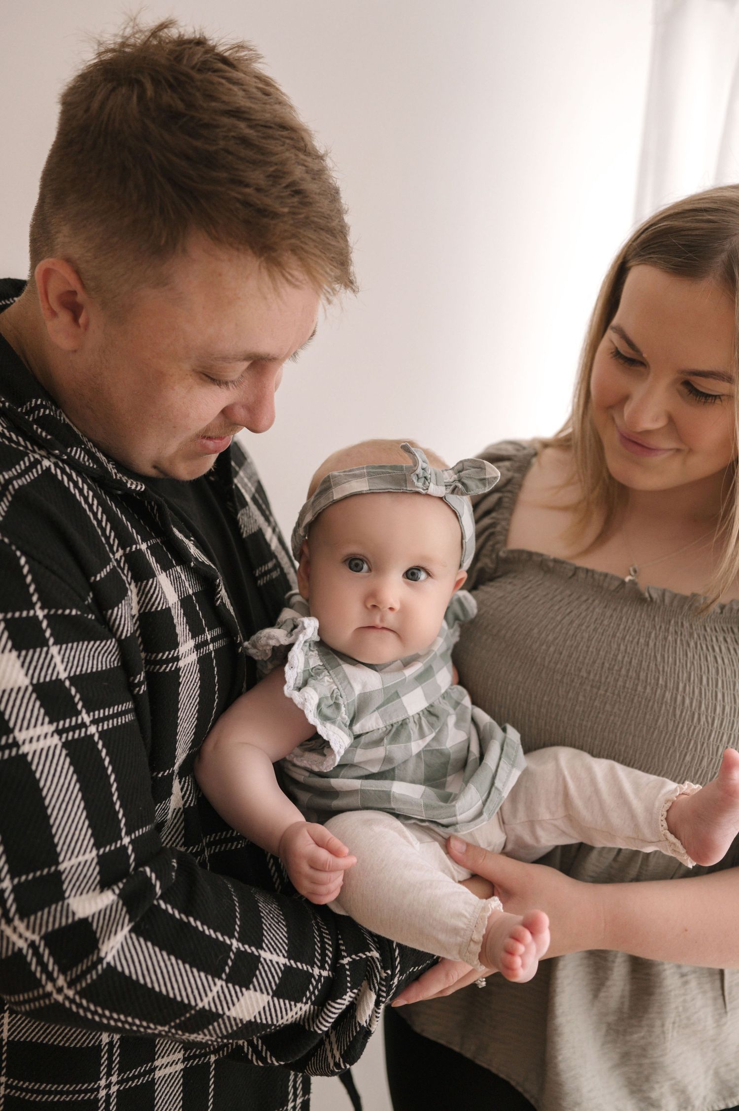 A young couple lovingly holds their baby girl dressed in a gray outfit with a bow headband.