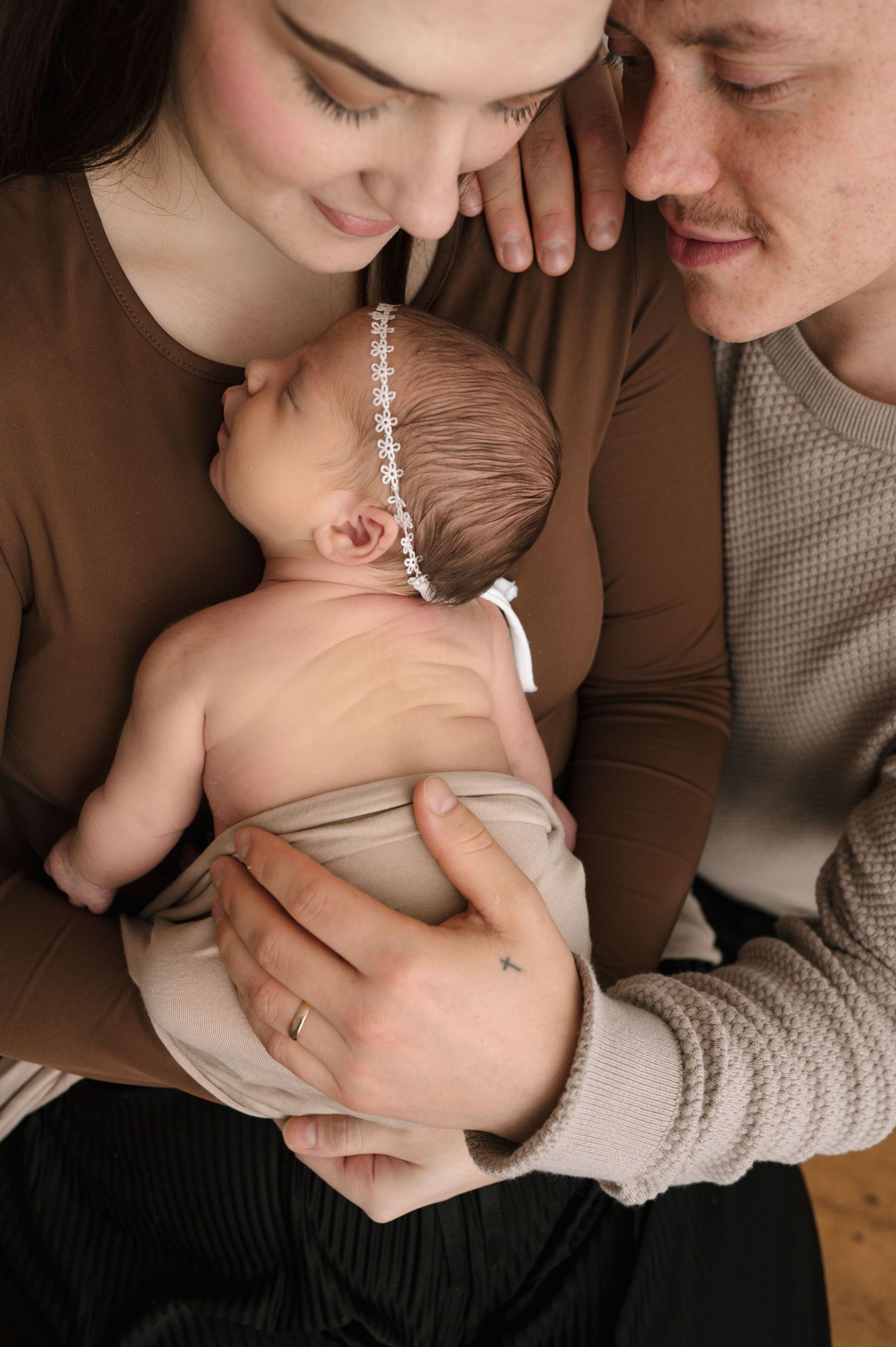 A couple lovingly gazes at their newborn baby girl wearing a delicate floral headband, wrapped in a soft beige swaddle.