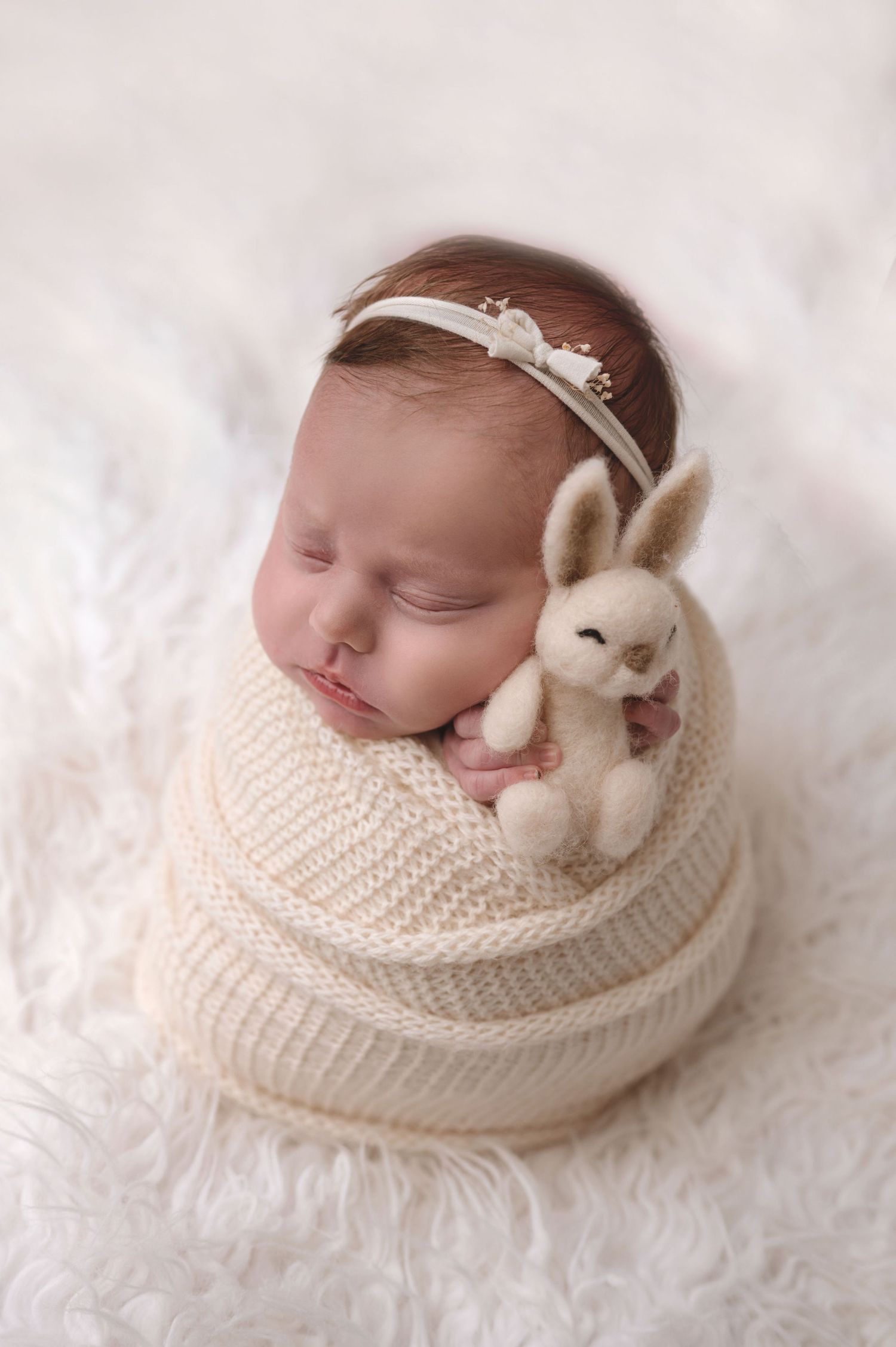 A sleeping newborn wrapped in cream knit fabric with a small plush bunny and delicate headband on soft white fur backdrop.