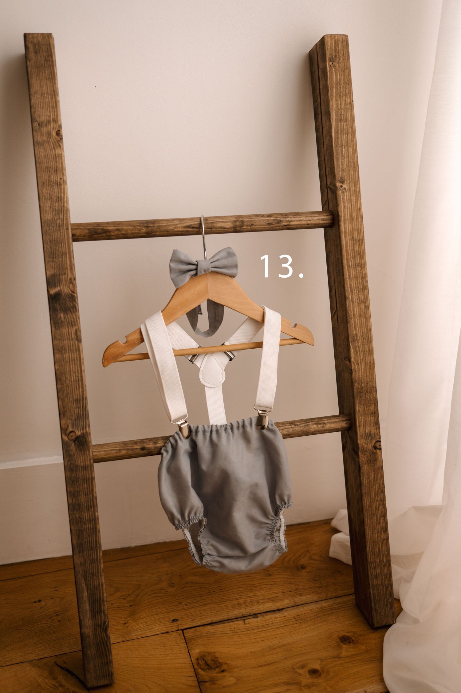 Four photos showing a gray baby romper with suspenders and bow tie displayed on wooden hangers against rustic ladder decor.