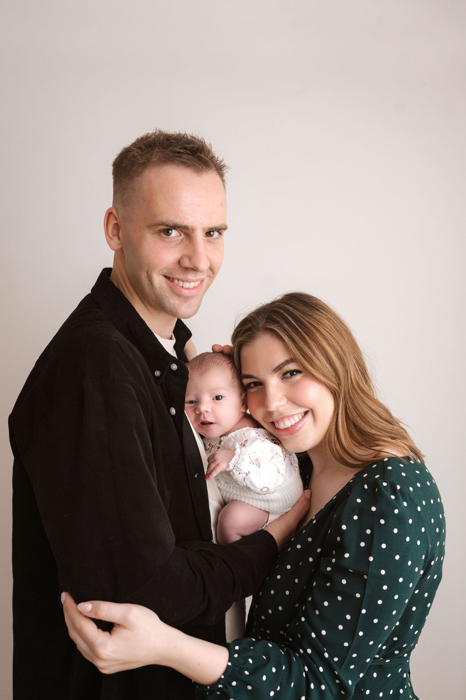 A young couple smiling and holding their newborn baby in a studio portrait photo.