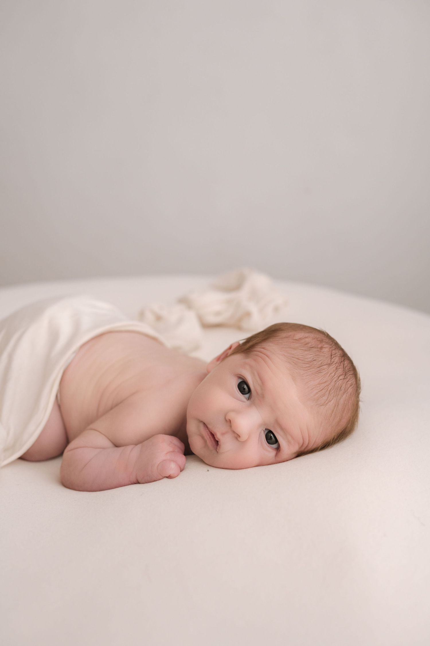 A newborn baby lies on a white surface wrapped in a soft cream blanket, gazing gently at the camera.