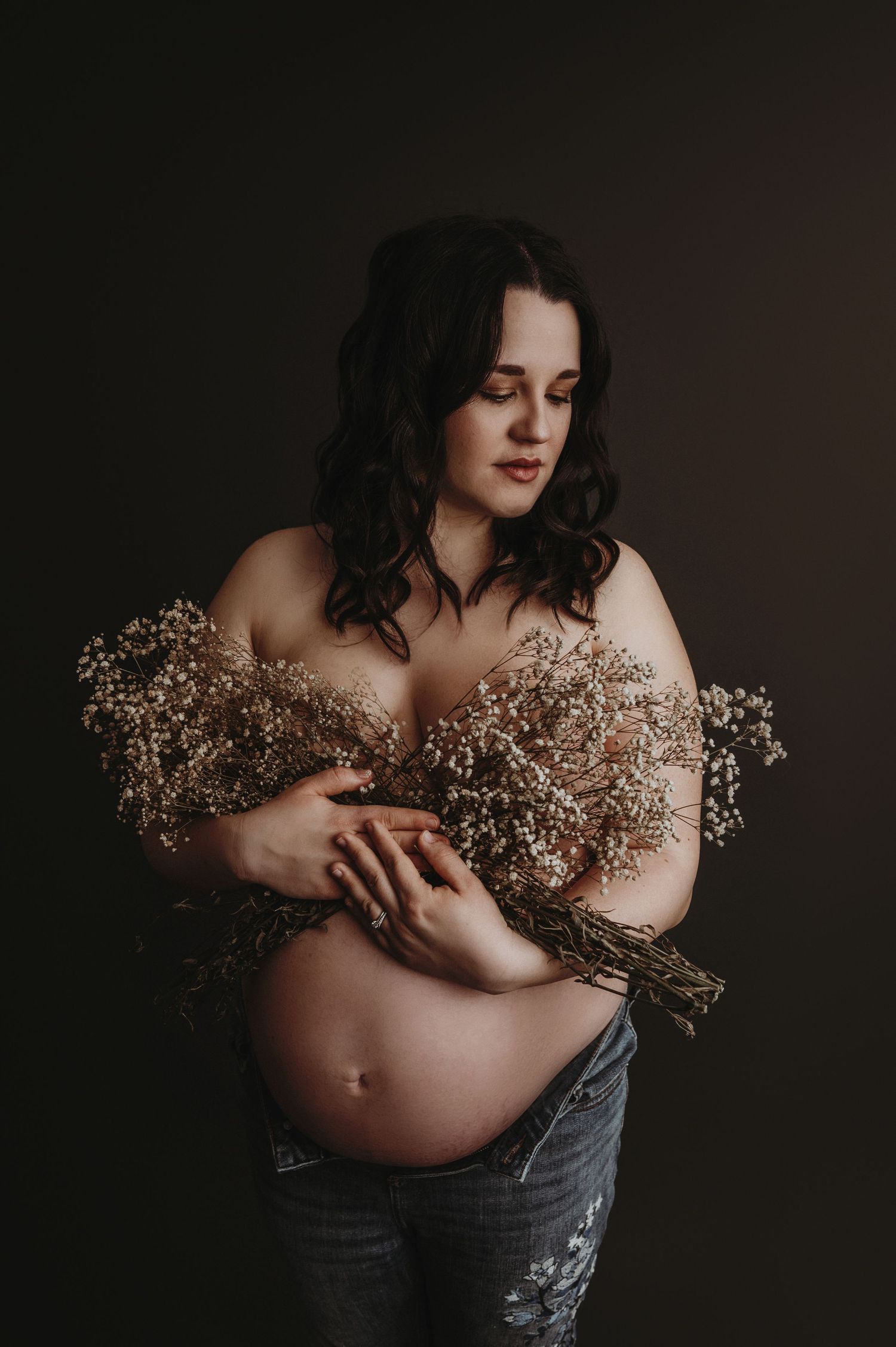 Artistic maternity photo showing an expectant mother holding dried flowers against a dark background.