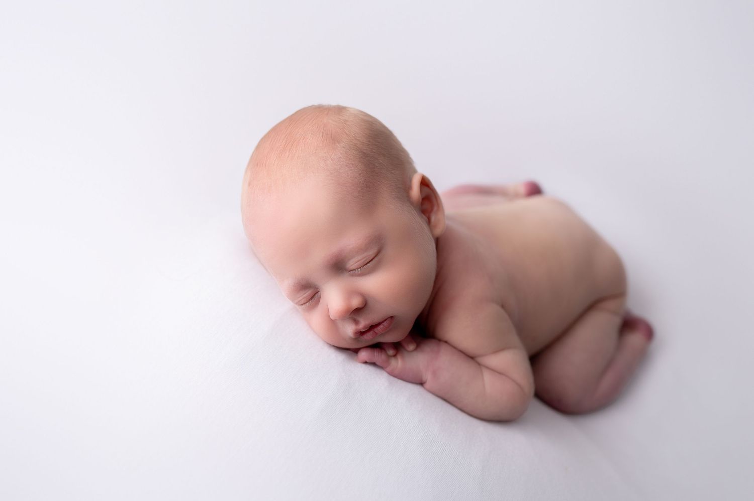 A newborn baby lies sleeping peacefully in a curled up position on a white background.