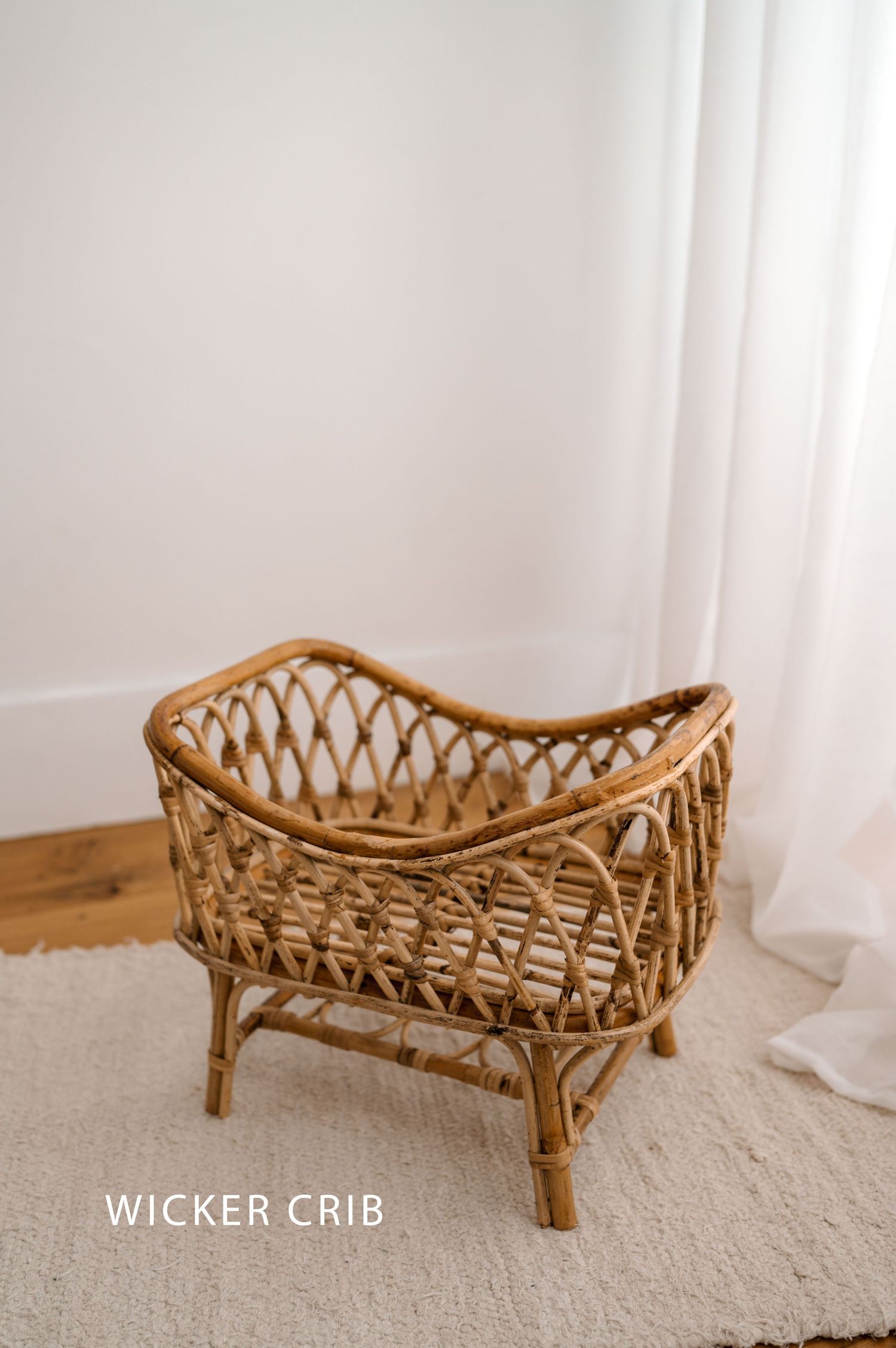 Vintage style natural wicker baby crib with curved design sits on beige carpet against white wall and curtain.