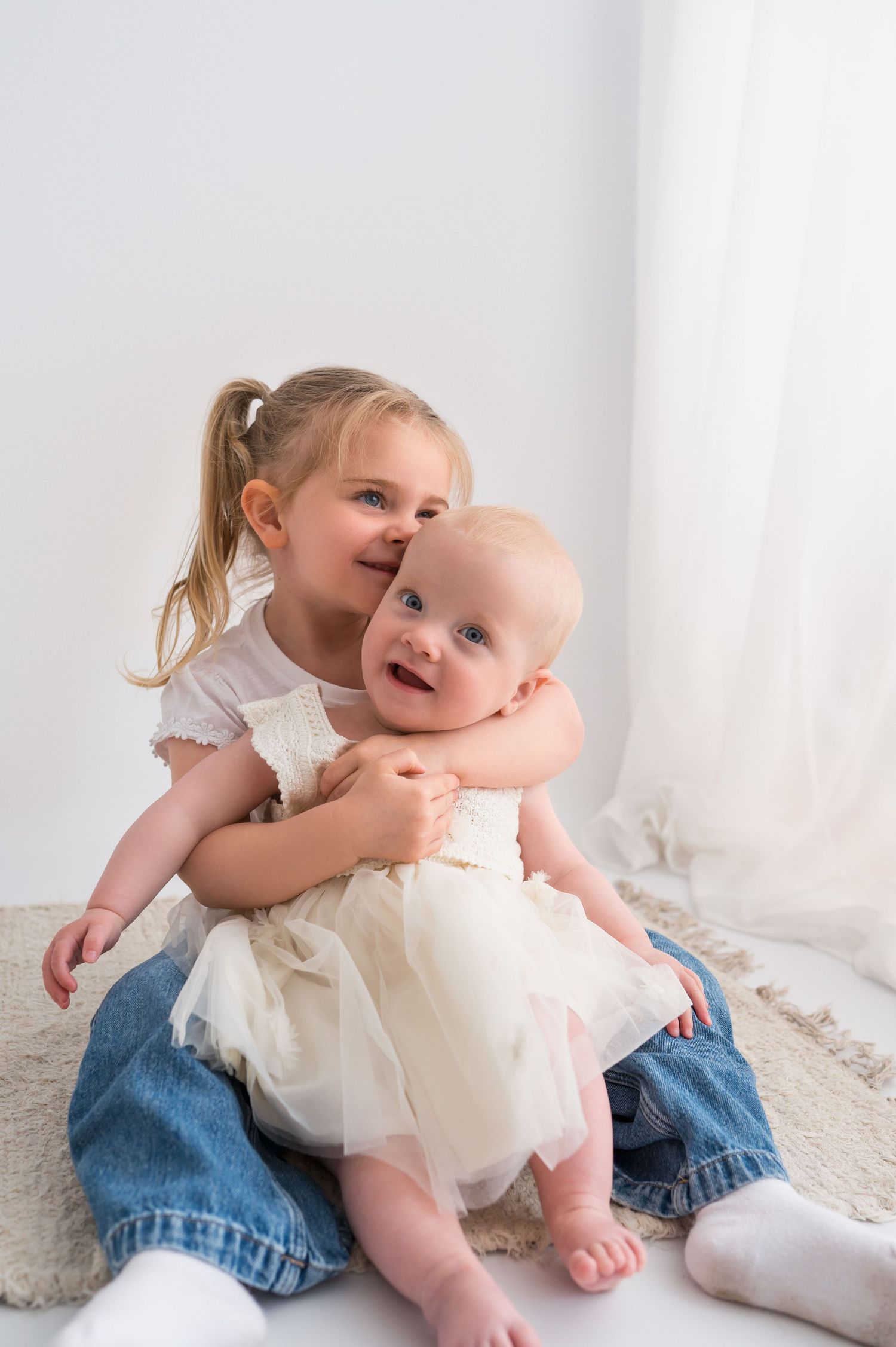 Young girl in jeans hugs smiling baby in white dress, both sitting on floor against white background.