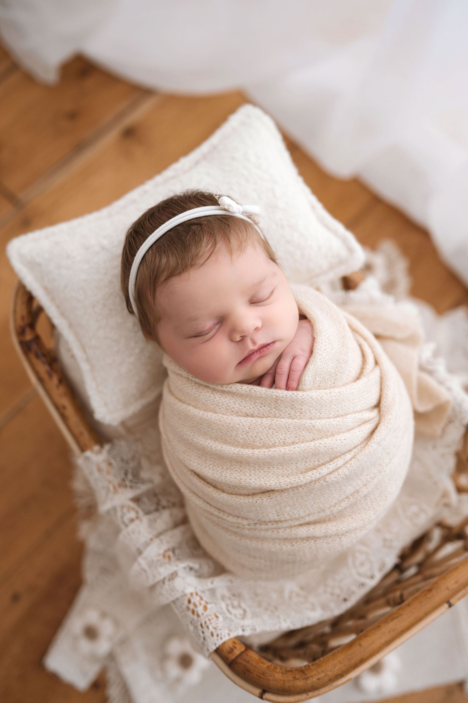 Newborn baby girl swaddled in cream wrap, sleeping peacefully in a wicker basket with a white pillow and headband.