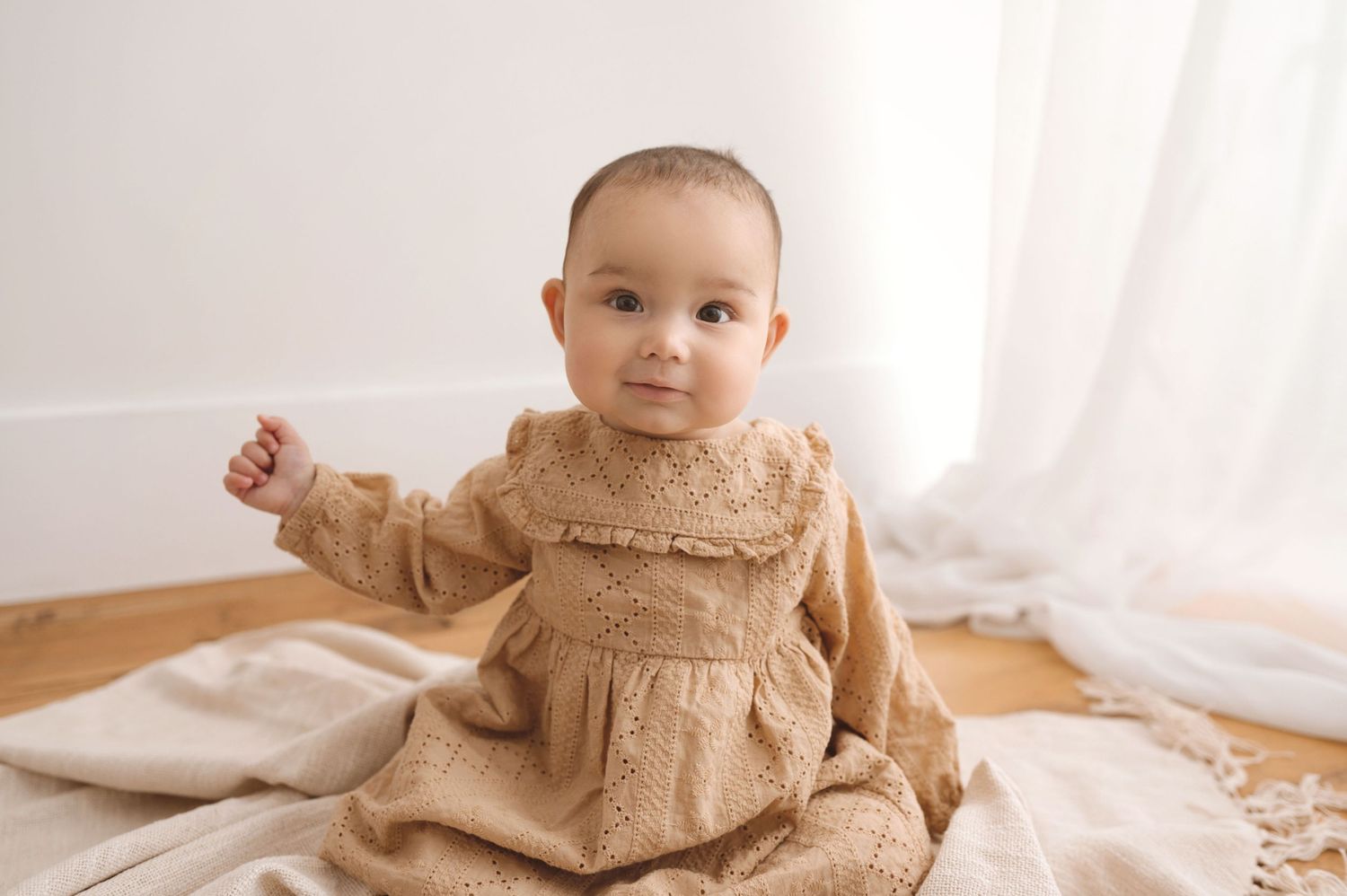 Small child sits on textured blanket wearing a beige ruffled dress while playing with scattered decorative elements.