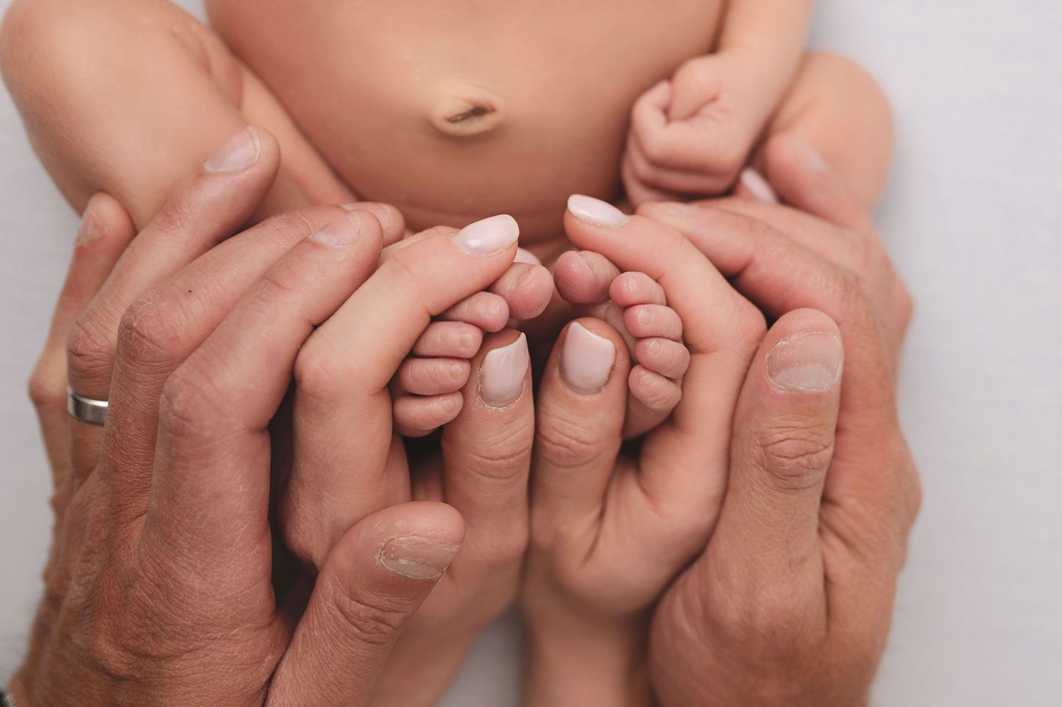 Adult hands gently cradle and support tiny baby feet and toes against a soft neutral background.