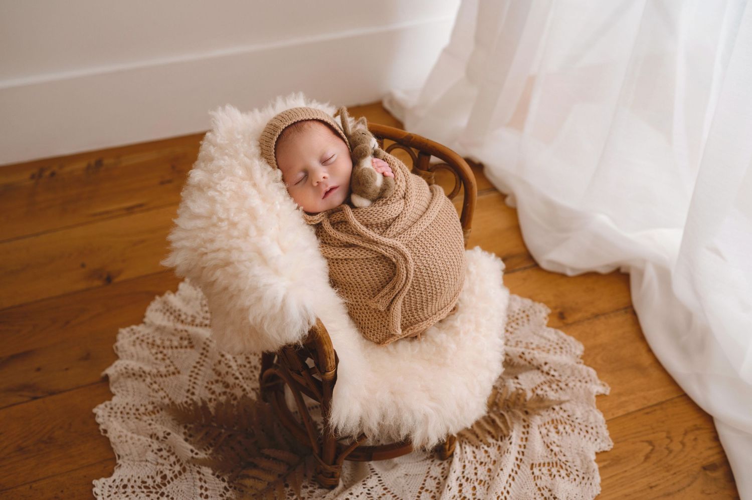 Sweet newborn sleeping on soft fluffy chair with lace doily on wood floor beside white curtain.