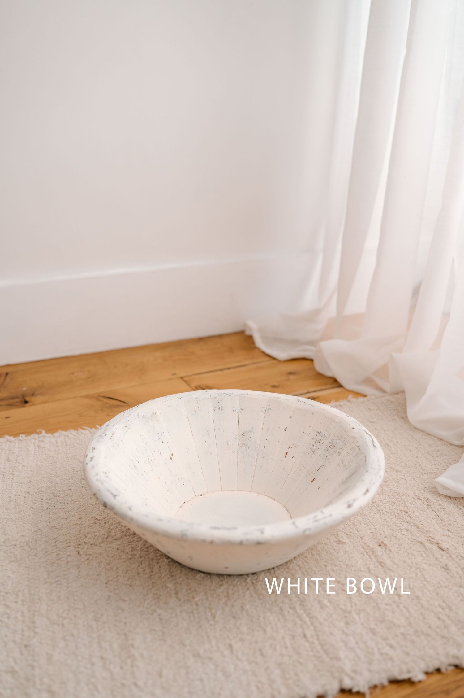 A simple white ceramic bowl with distressed finish rests on a cream colored rug near flowing white curtains.