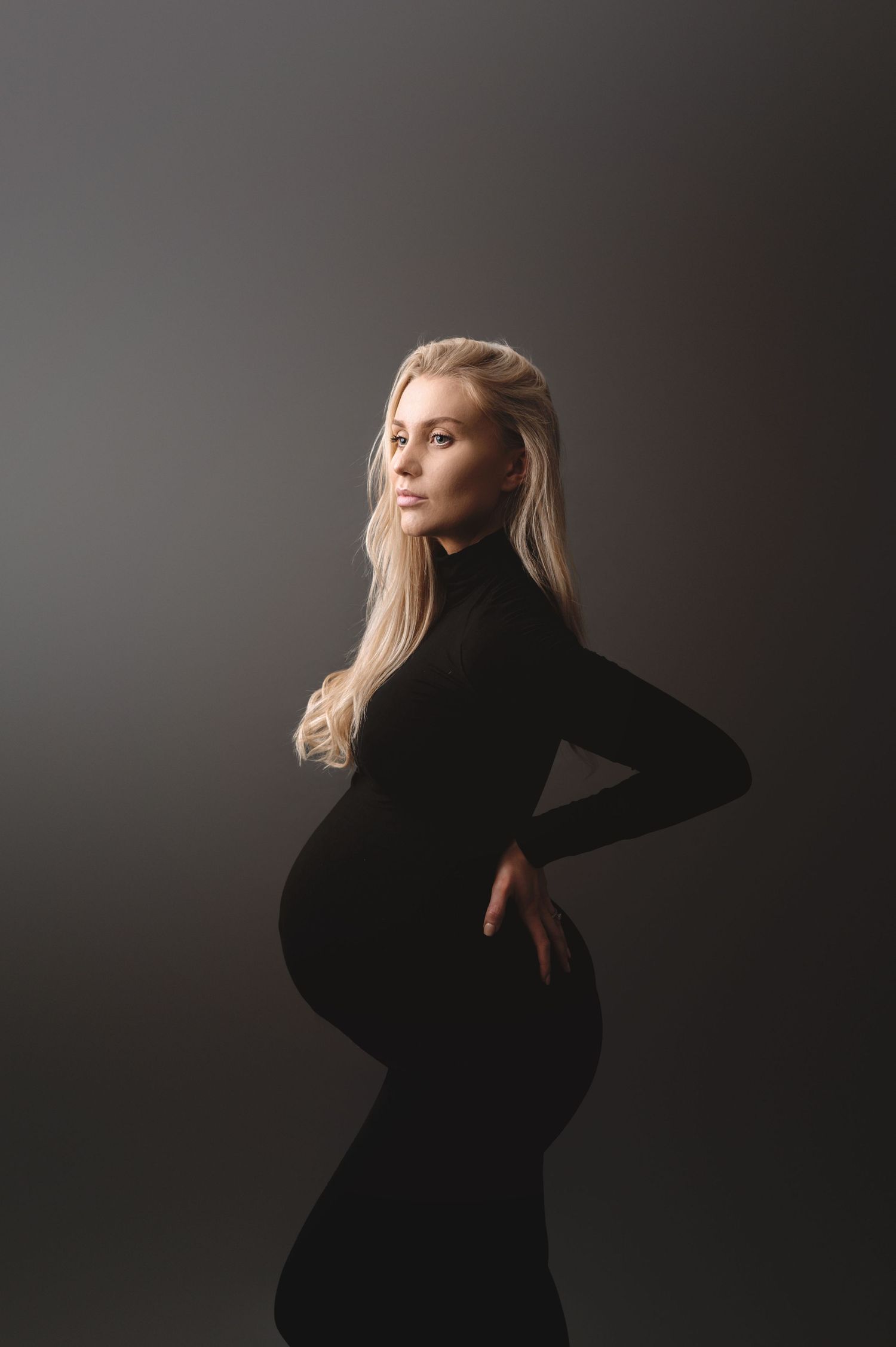 A silhouette stands in dramatic lighting wearing a fitted black gown against a dark background.