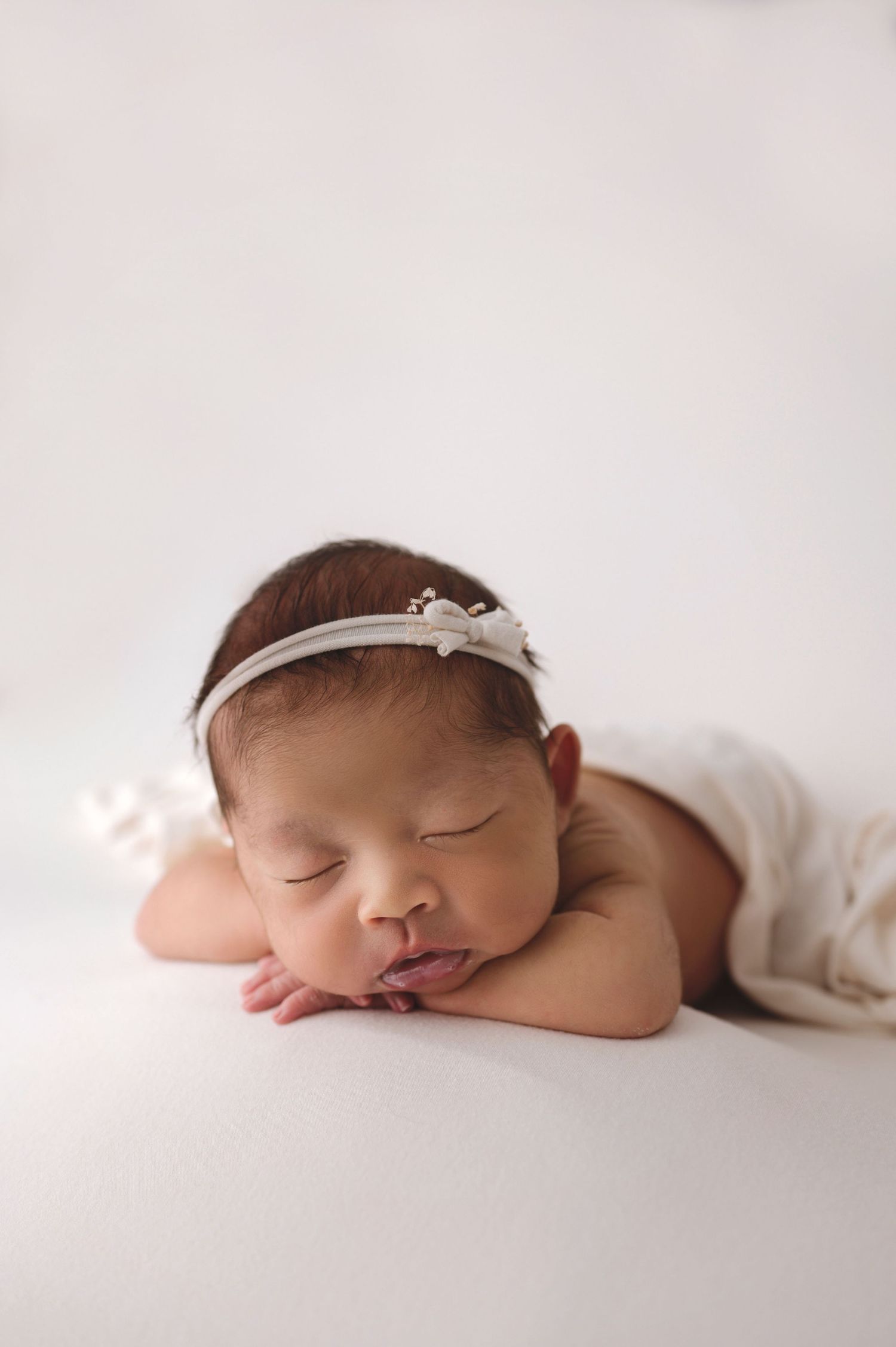 Peaceful sleeping newborn in white wrap and delicate headband rests on light backdrop during professional photo session.