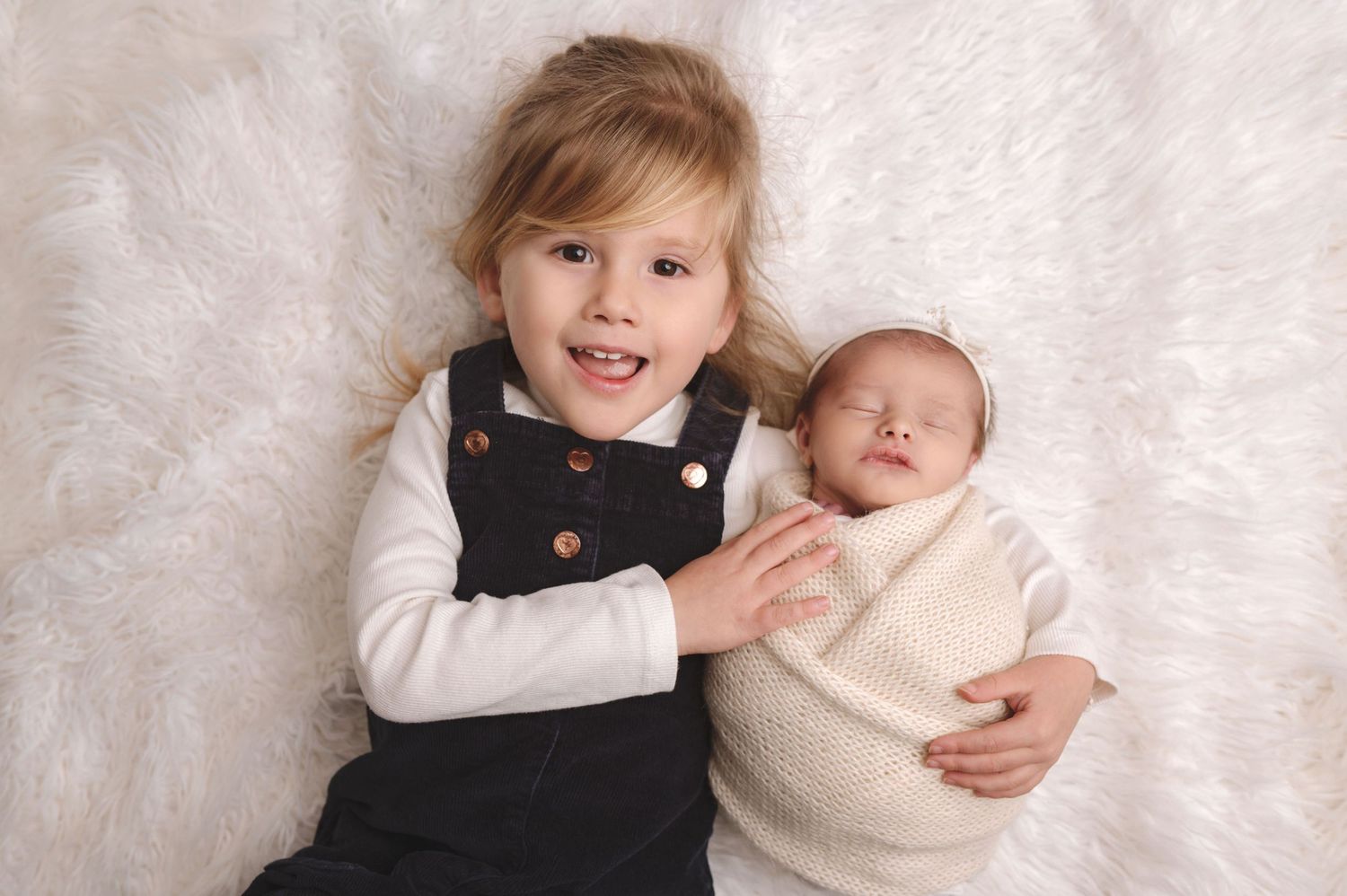 Young child in black dress laying on white fur backdrop with swaddled sleeping baby.