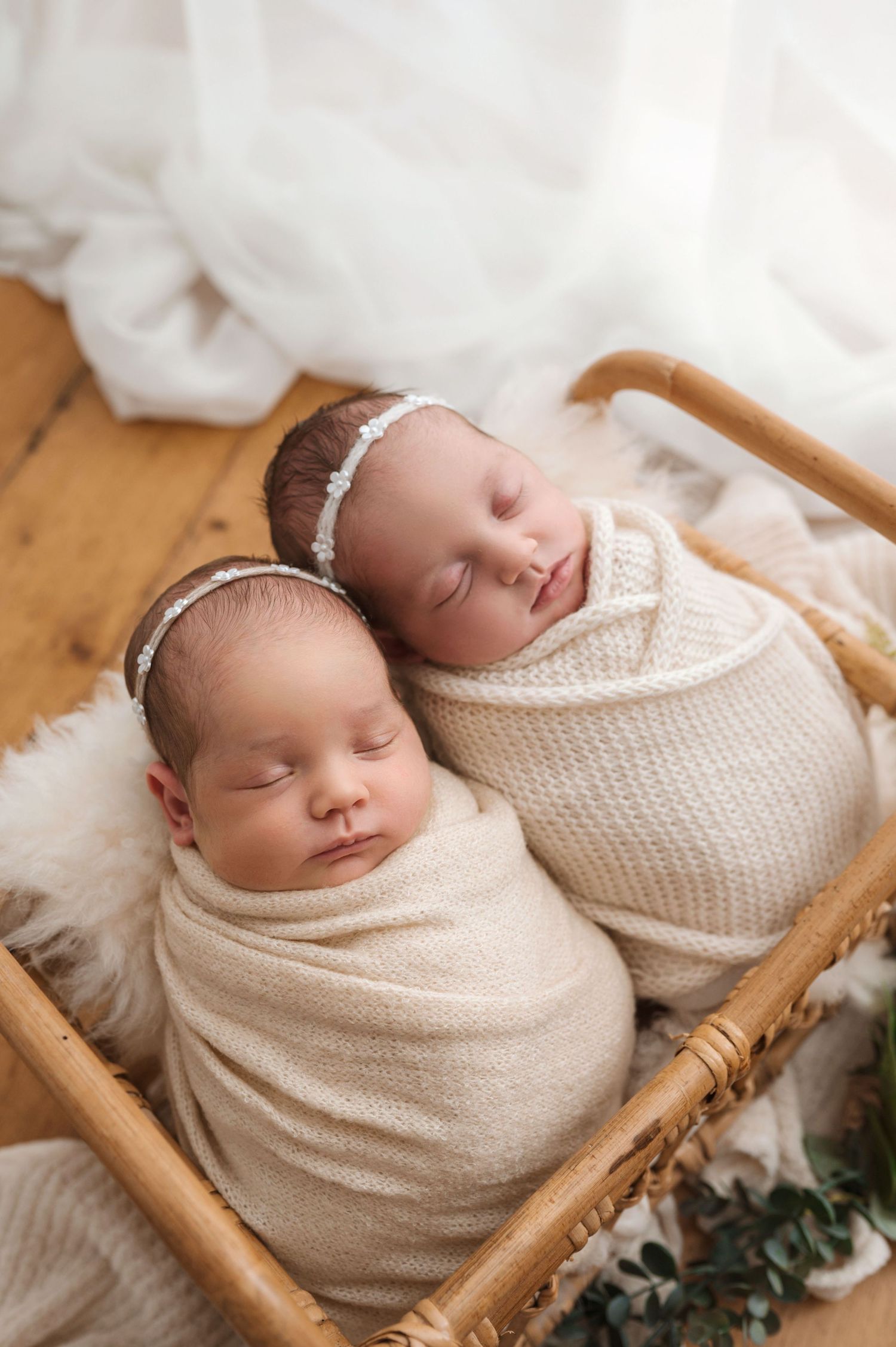 Two swaddled newborn twins sleeping peacefully in a wicker basket with soft cream wraps and a delicate headband.