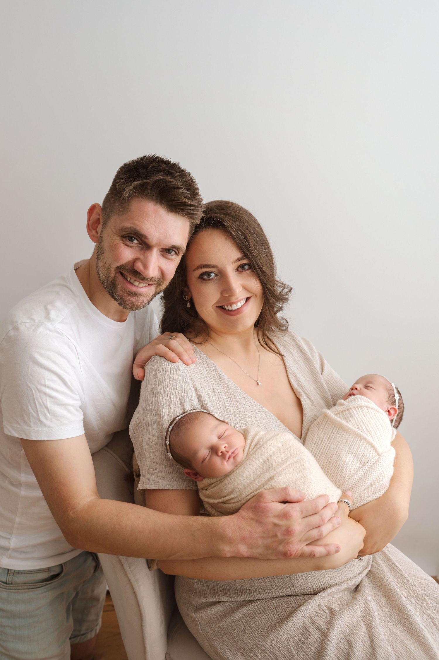 A couple in light-colored clothes holds their twin newborn babies wrapped in cream-colored swaddles against a neutral background.