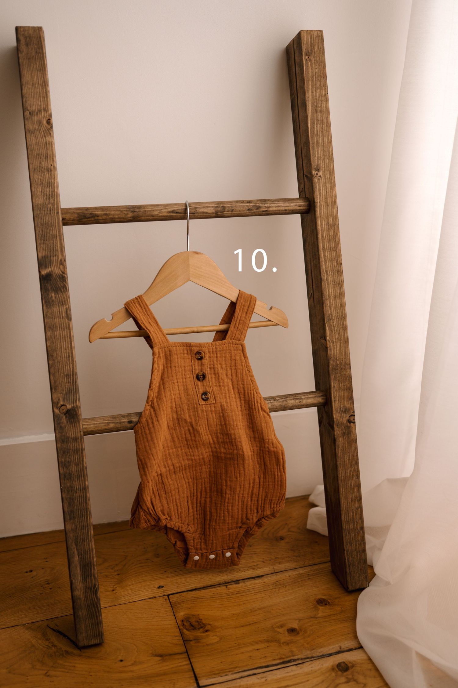 Brown muslin baby romper displayed on wooden hanger against rustic ladder and white wall.