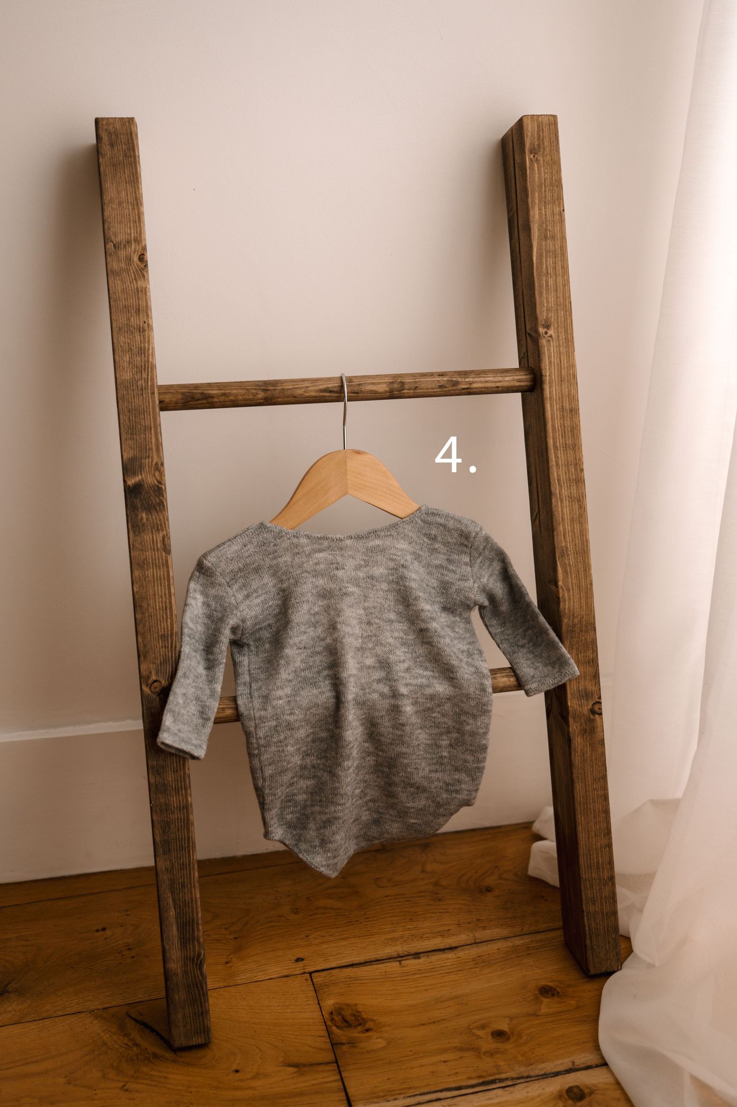 A gray sweater hanging on a wooden ladder shelf against a white wall and wooden floor.