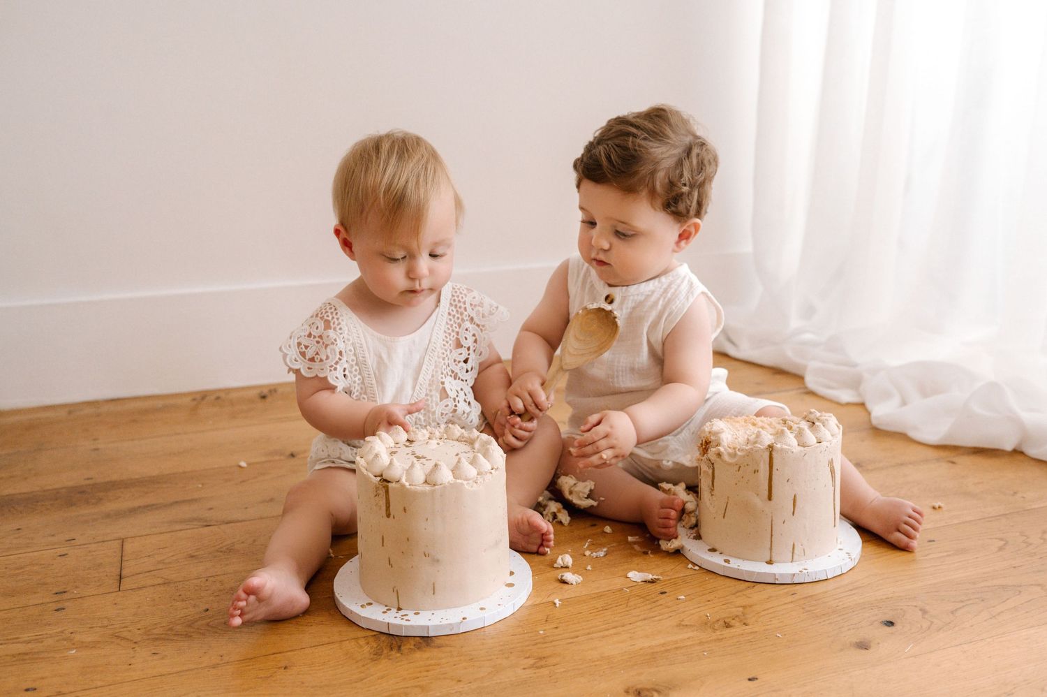 Two children in pink outfits sit on wooden floor with matching cream-colored cakes during a cake smash photoshoot.