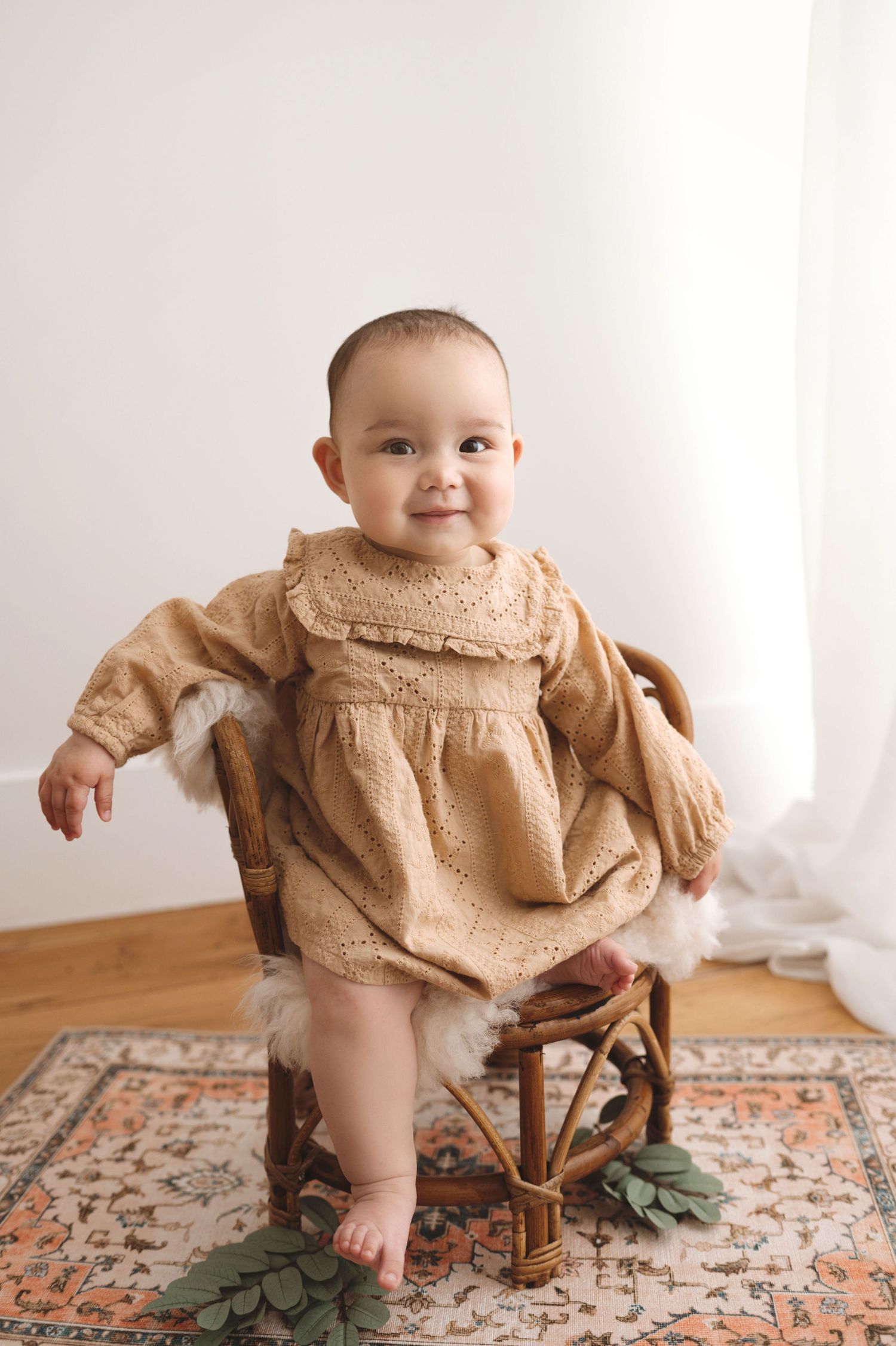 Small child wearing tan vintage-style dress sits on wooden chair atop floral rug in minimalist bright room.
