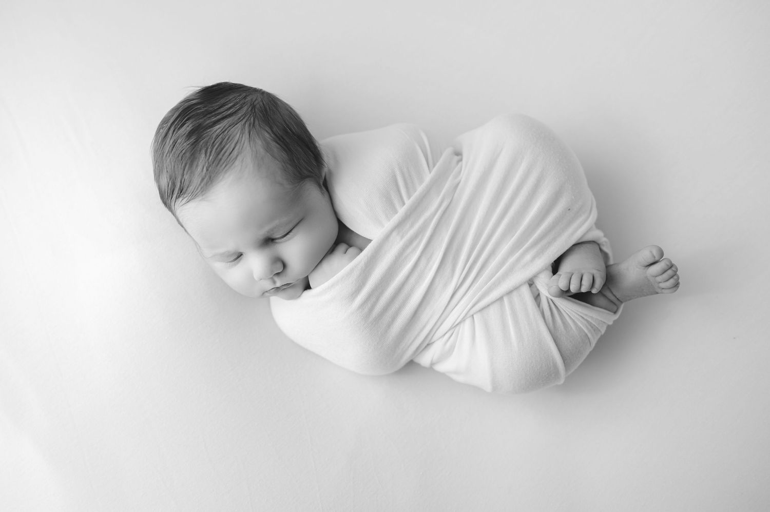 A newborn baby rests peacefully wrapped in white cloth against a light background in black and white photography.