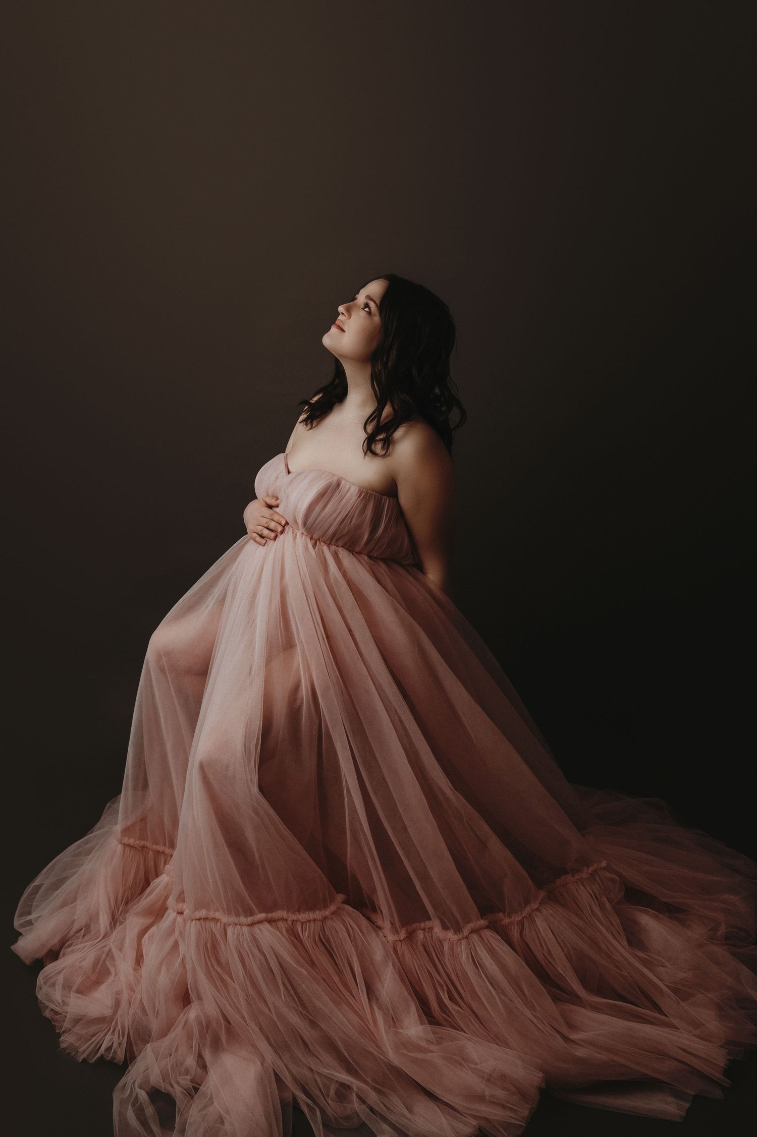 A dramatic maternity portrait features a flowing pink tulle gown against a dark background in moody lighting.