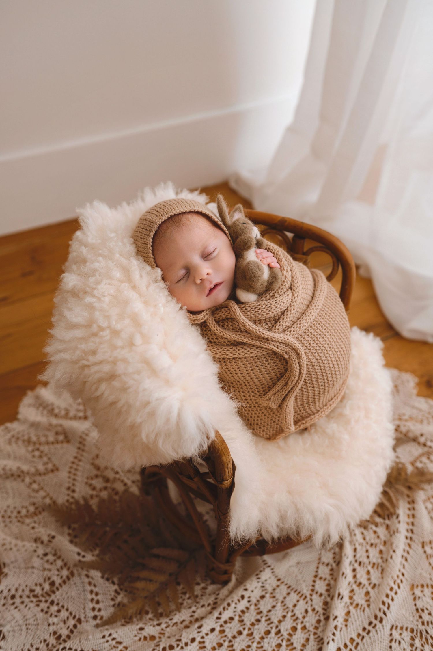A sleeping newborn swaddled in a brown knit blanket rests on a delicate white lace surface near sheer curtains.