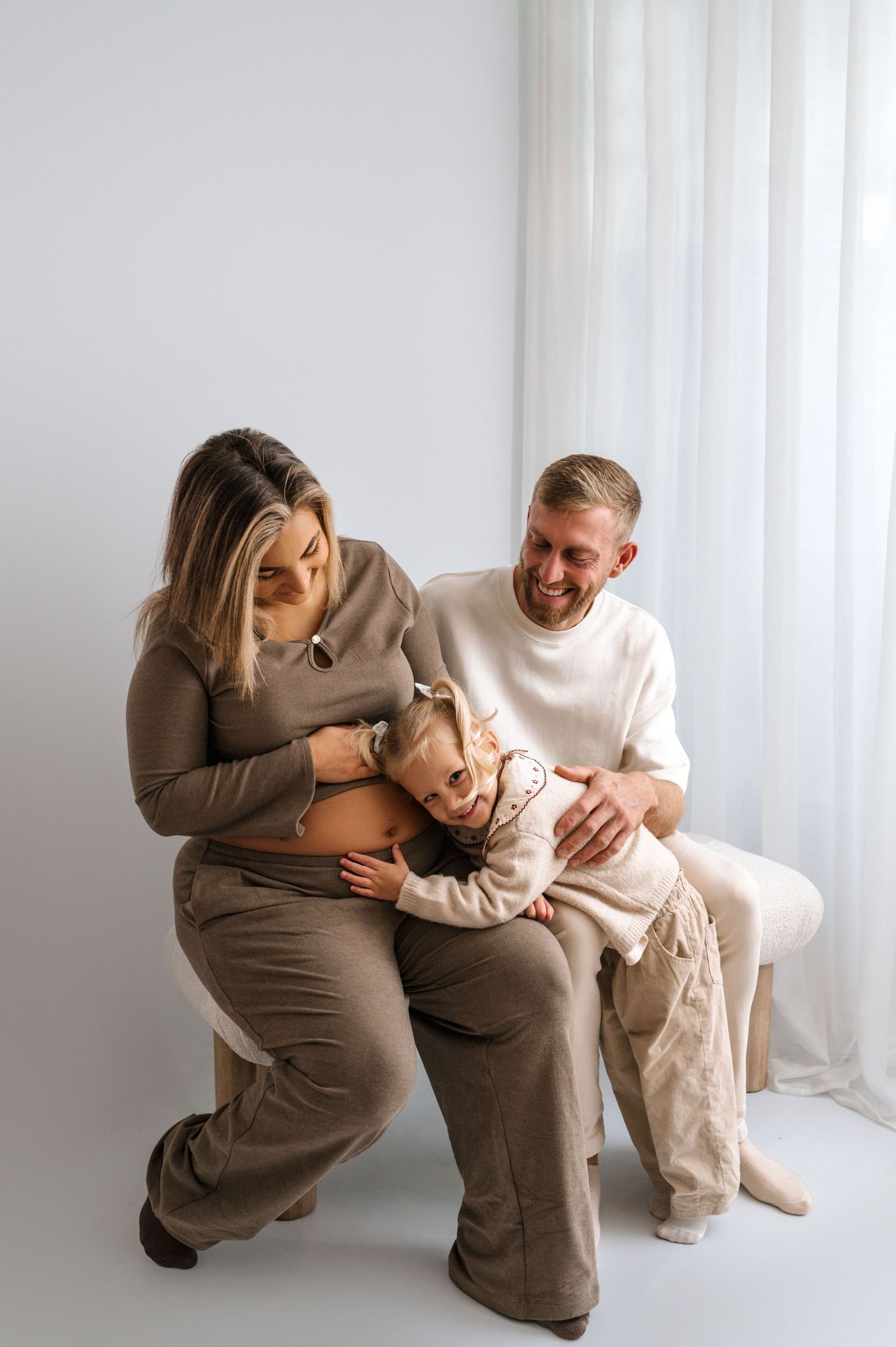 Pregnant woman and man smiling with young child hugging baby bump in bright white studio setting.