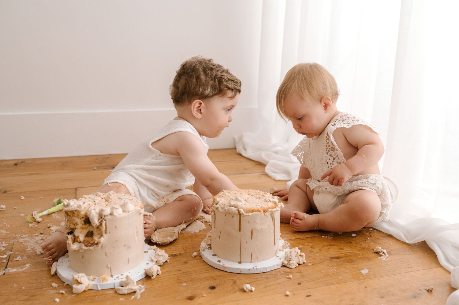Two children in matching white outfits sit together on a hardwood floor enjoying birthday cakes.