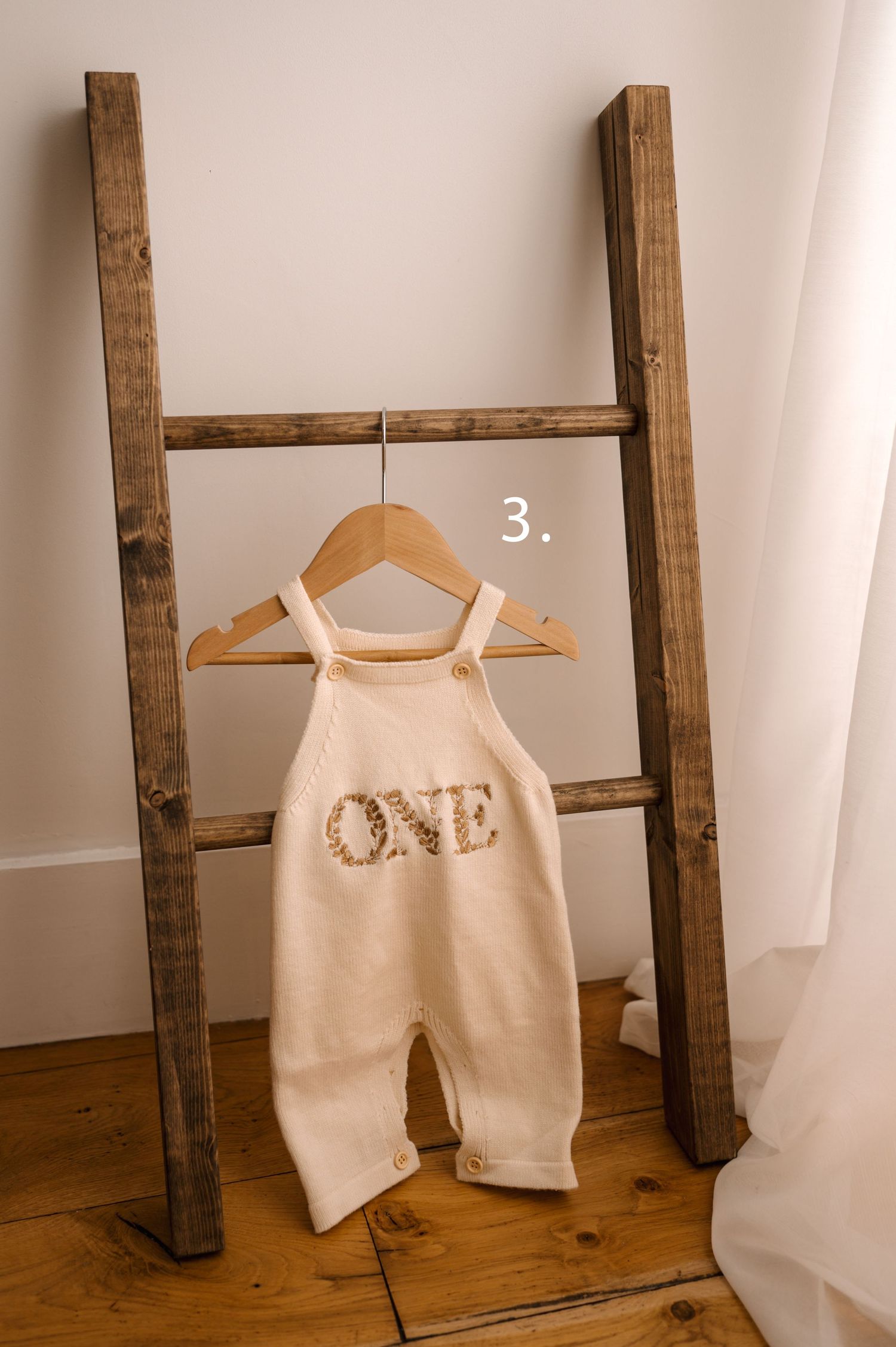 Cream colored baby romper hanging on wooden hanger against rustic ladder display with white curtain backdrop.