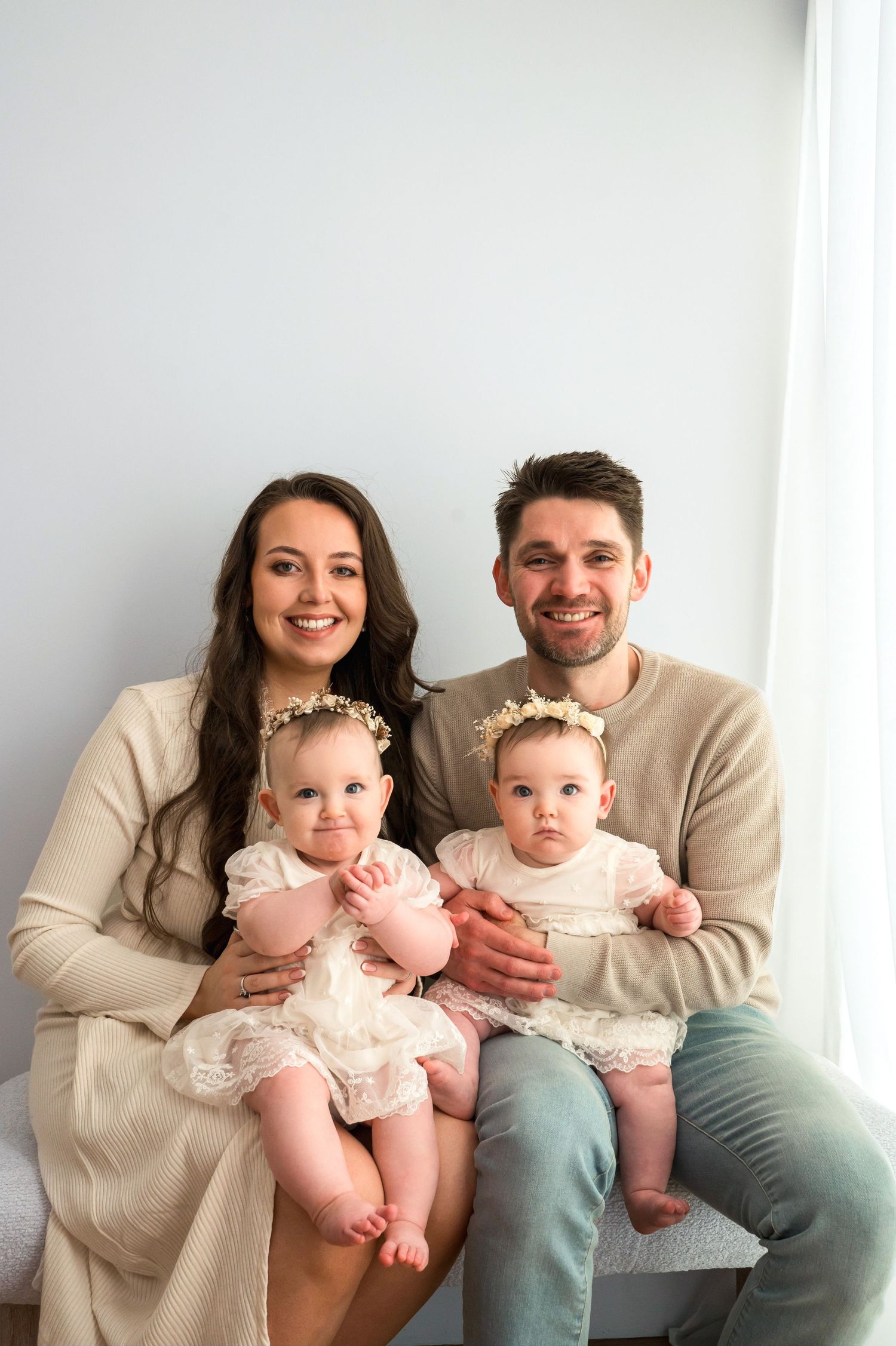 Smiling family portrait with parents holding twin baby girls in white lace dresses and floral crowns against a light background.