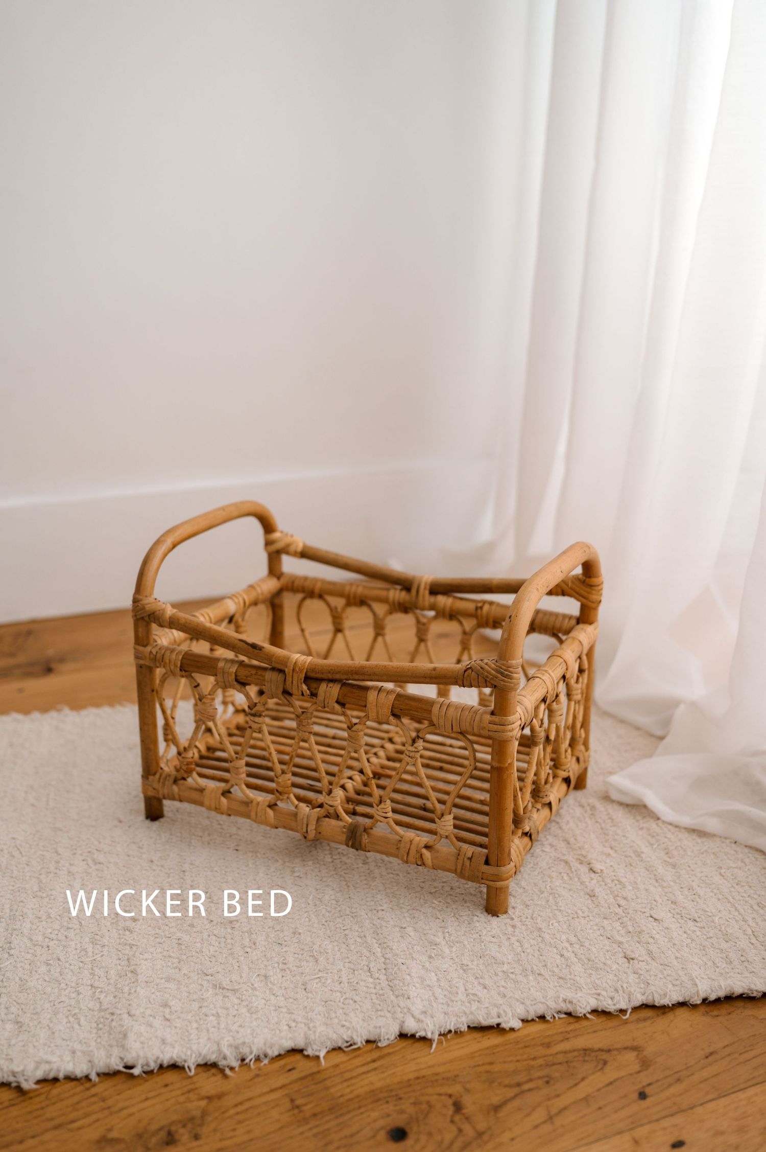 Small wicker doll bed or pet bed displayed on white carpet against light curtained wall.