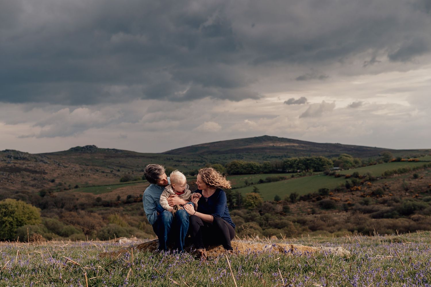 Two parents and a baby sit together in a wildflower meadow under dramatic stormy skies for a Dartmoor Family Photoshoot