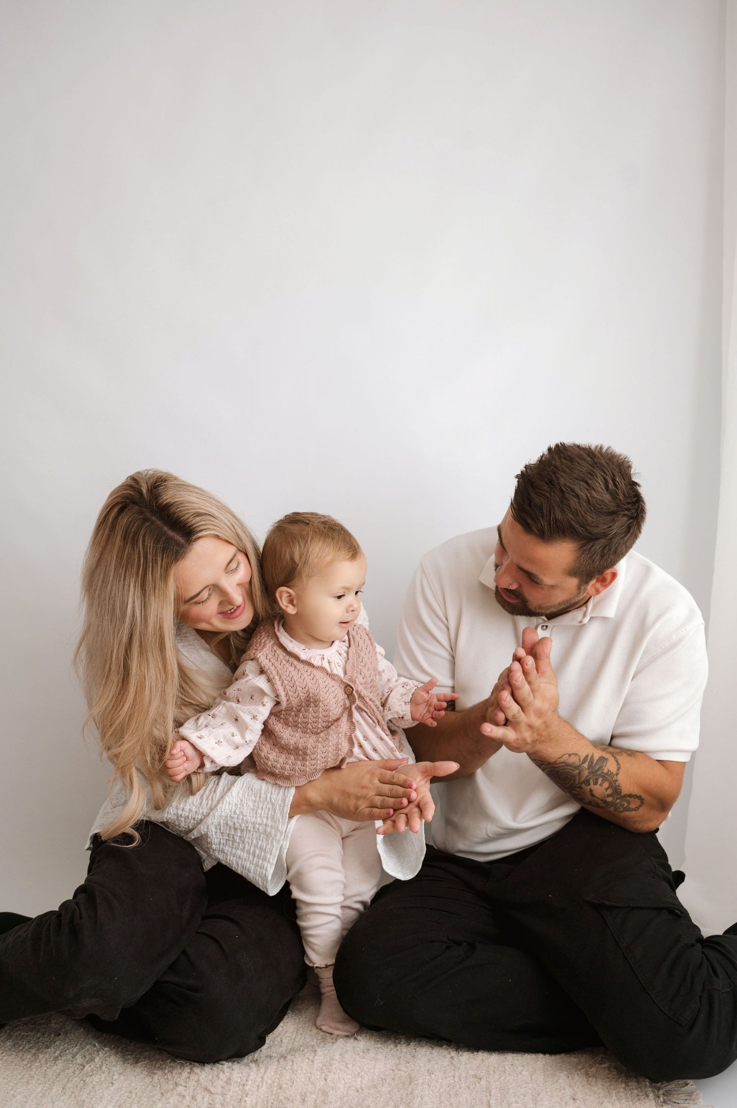 Young family with tattooed father, blonde mother, and baby girl sitting together on white floor smiling.