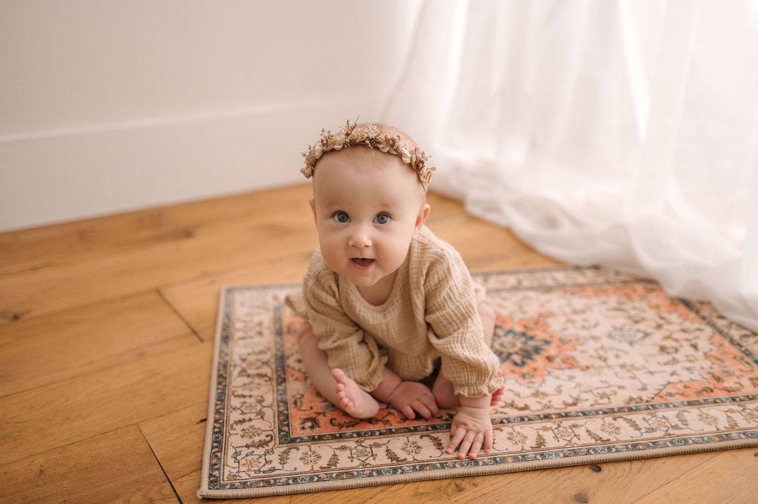 A baby wearing a floral crown crawls on a decorative area rug in a room with hardwood floors.