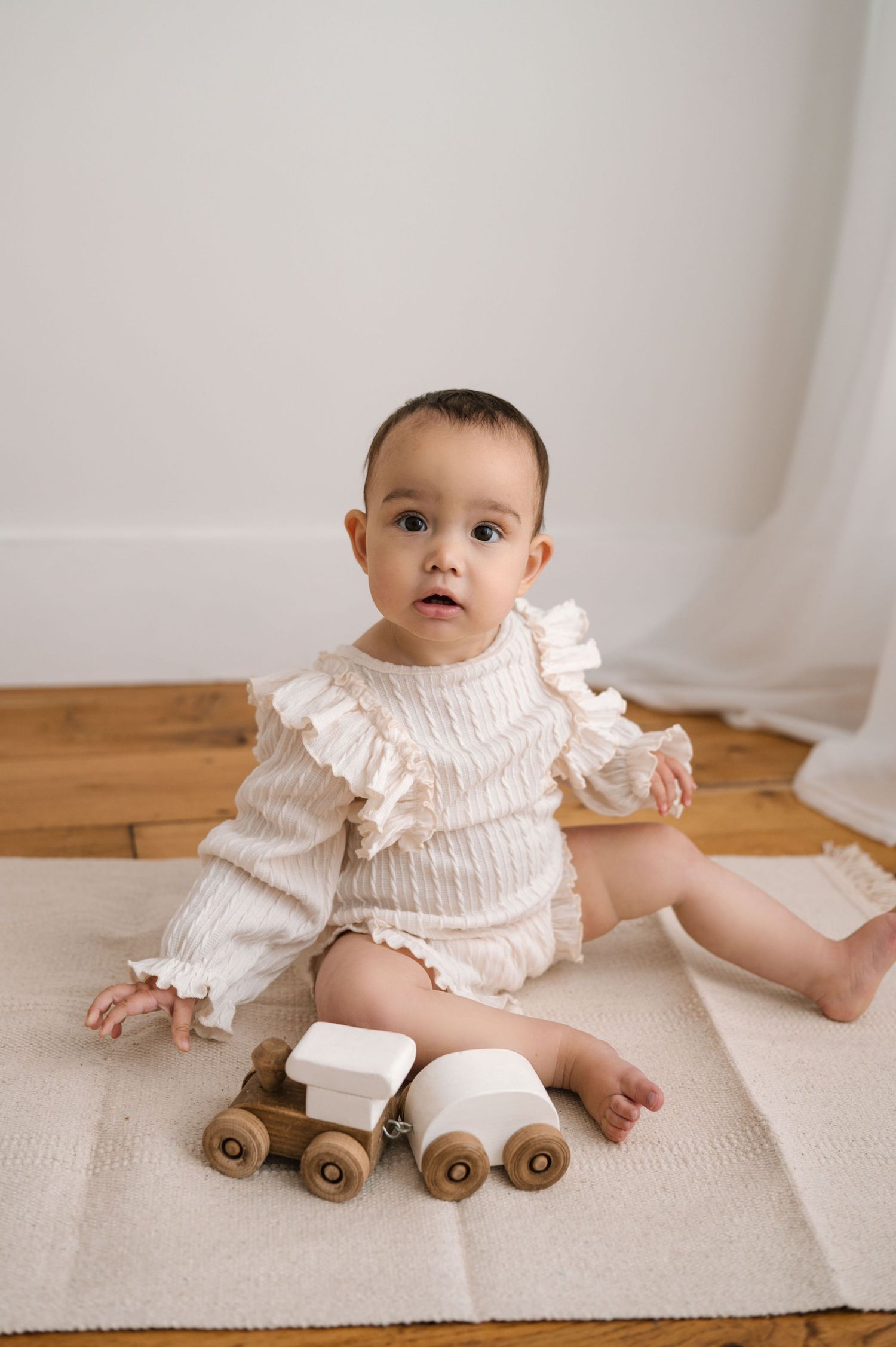 Baby in white ruffled outfit sits on cream rug with wooden toy train set.