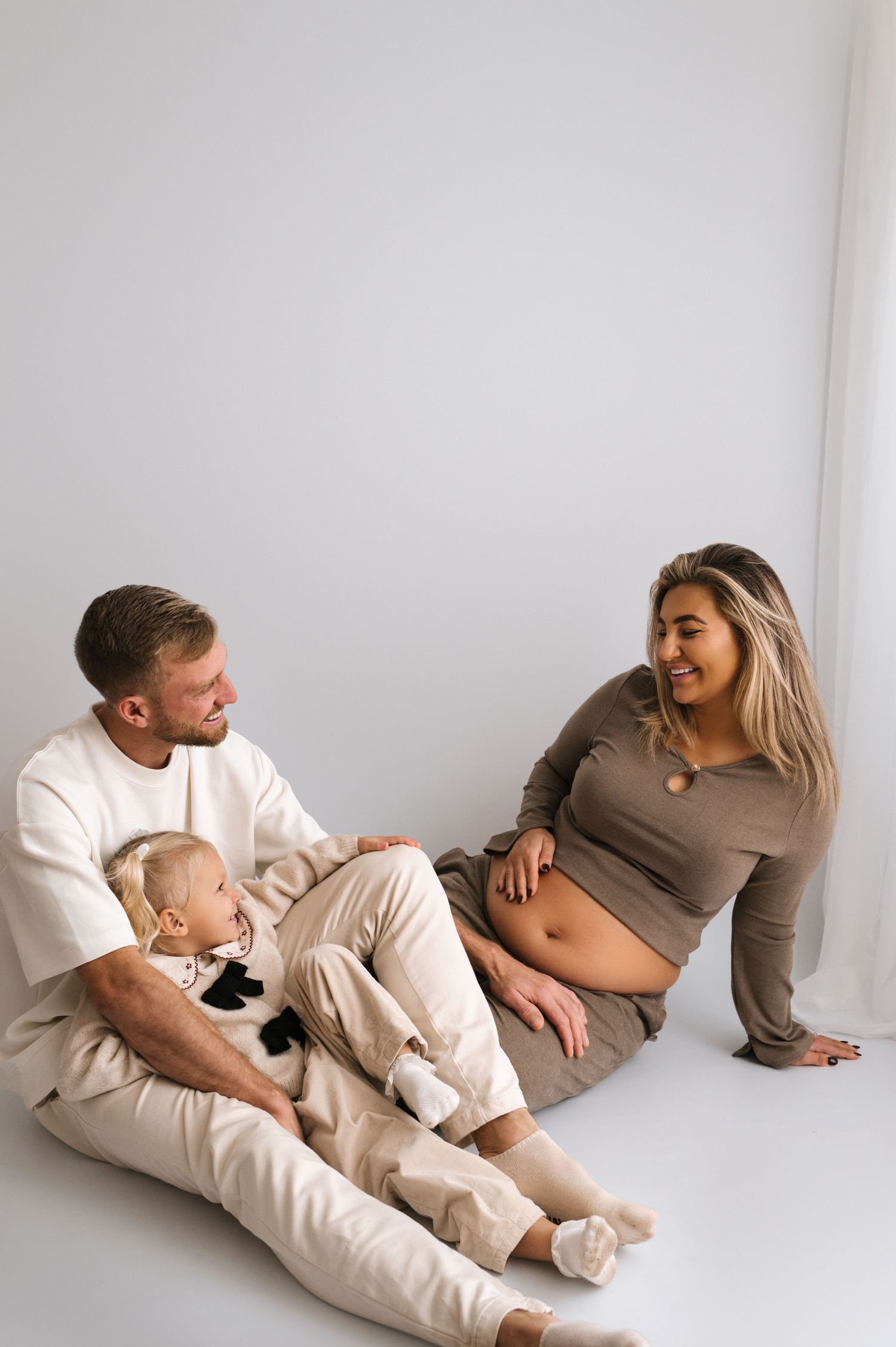 A happy family sits on a white floor, with a man holding a baby while a pregnant woman smiles beside them.