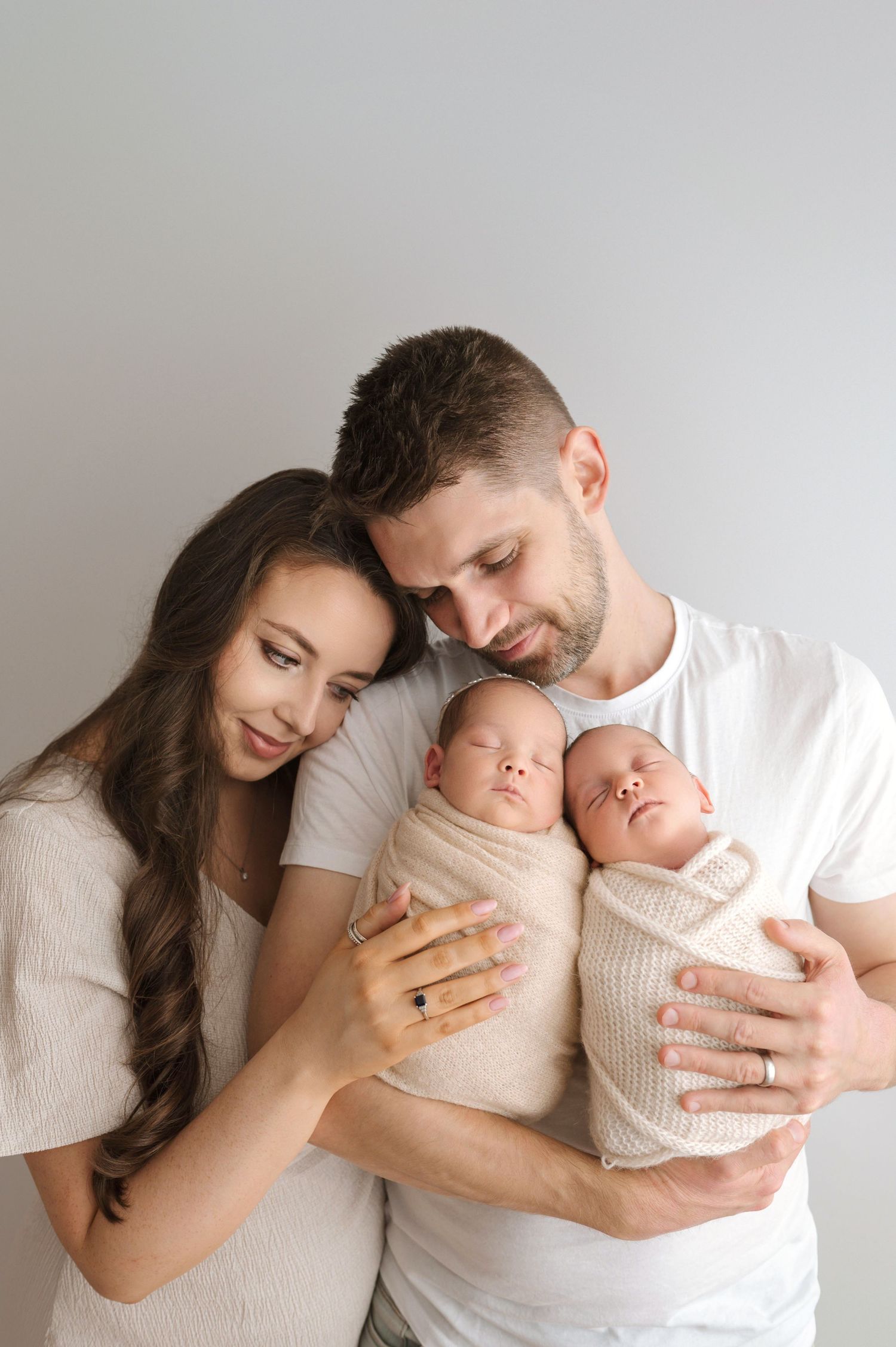 A young couple in white outfits tenderly holds their two sleeping newborn twins wrapped in beige swaddles.