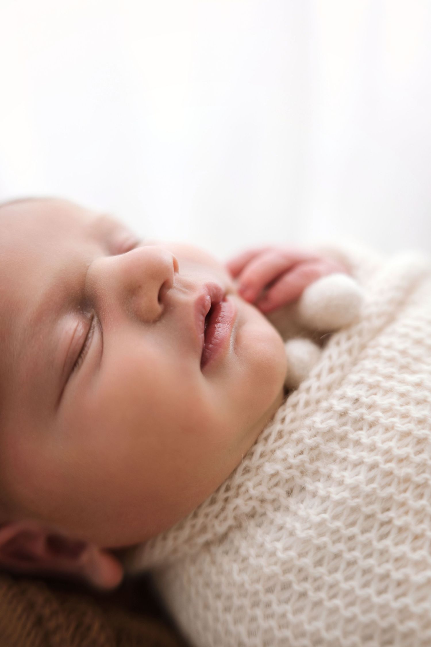 A peaceful newborn baby sleeping soundly, wrapped in a soft cream knit blanket with a tiny pompom detail.