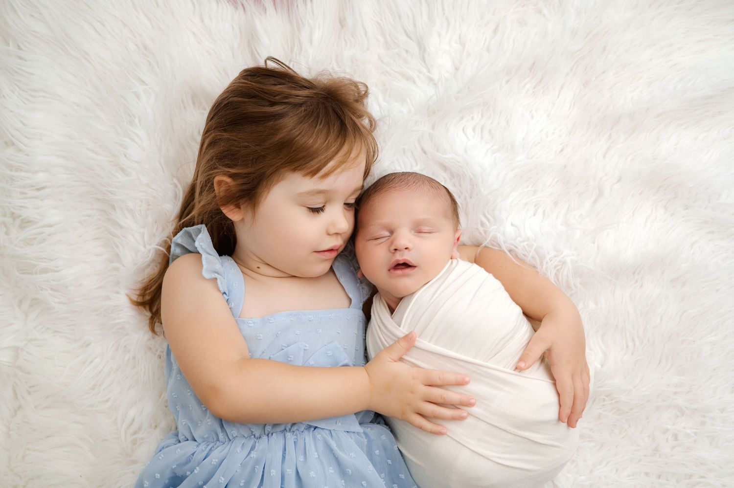 Young girl in blue dress tenderly holds sleeping swaddled newborn baby on white fluffy blanket.