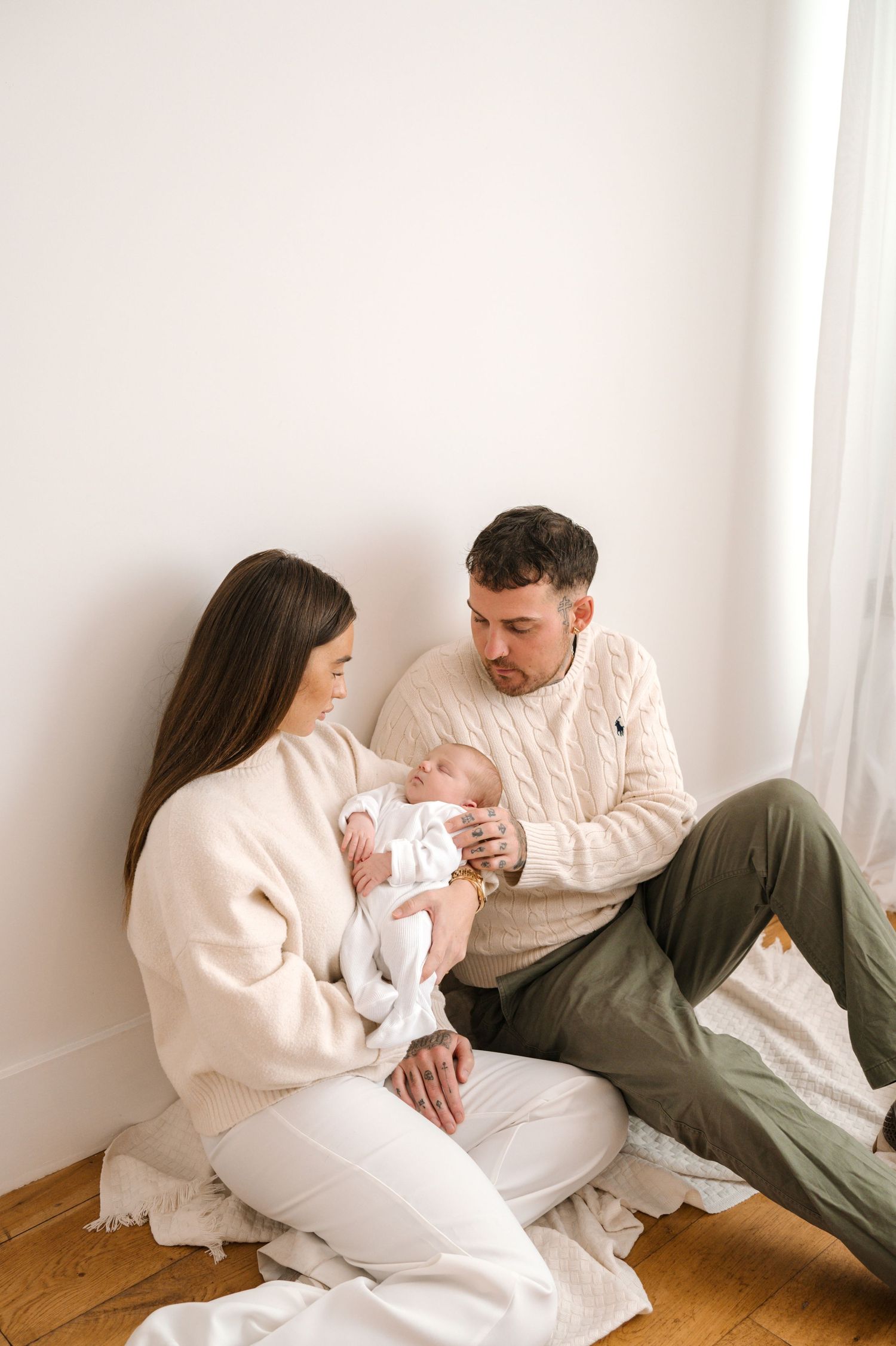 A couple sits on the floor cradling their newborn baby, dressed in cozy cream and white outfits in a bright minimalist room.